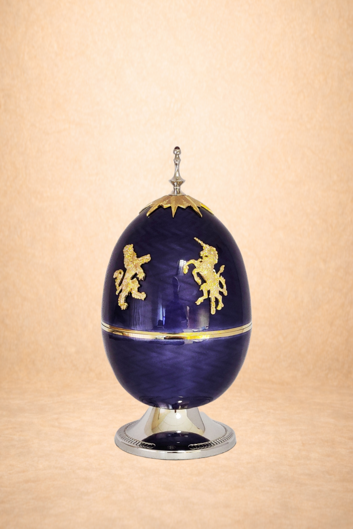 A dark blue, decorative egg-shaped object with gold lion and unicorn designs, topped with a gold starburst finial, set on a silver stand against a plain beige background.