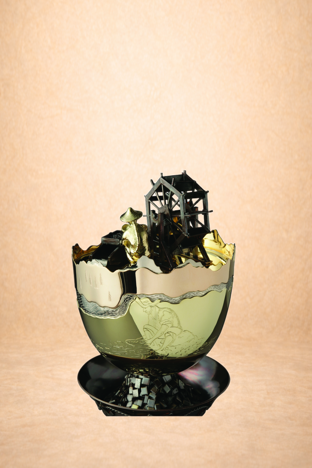 A decorative glass with engraved figures, topped with a miniature black watermill, a golden figure, and a golden scoop, set against a plain beige background.