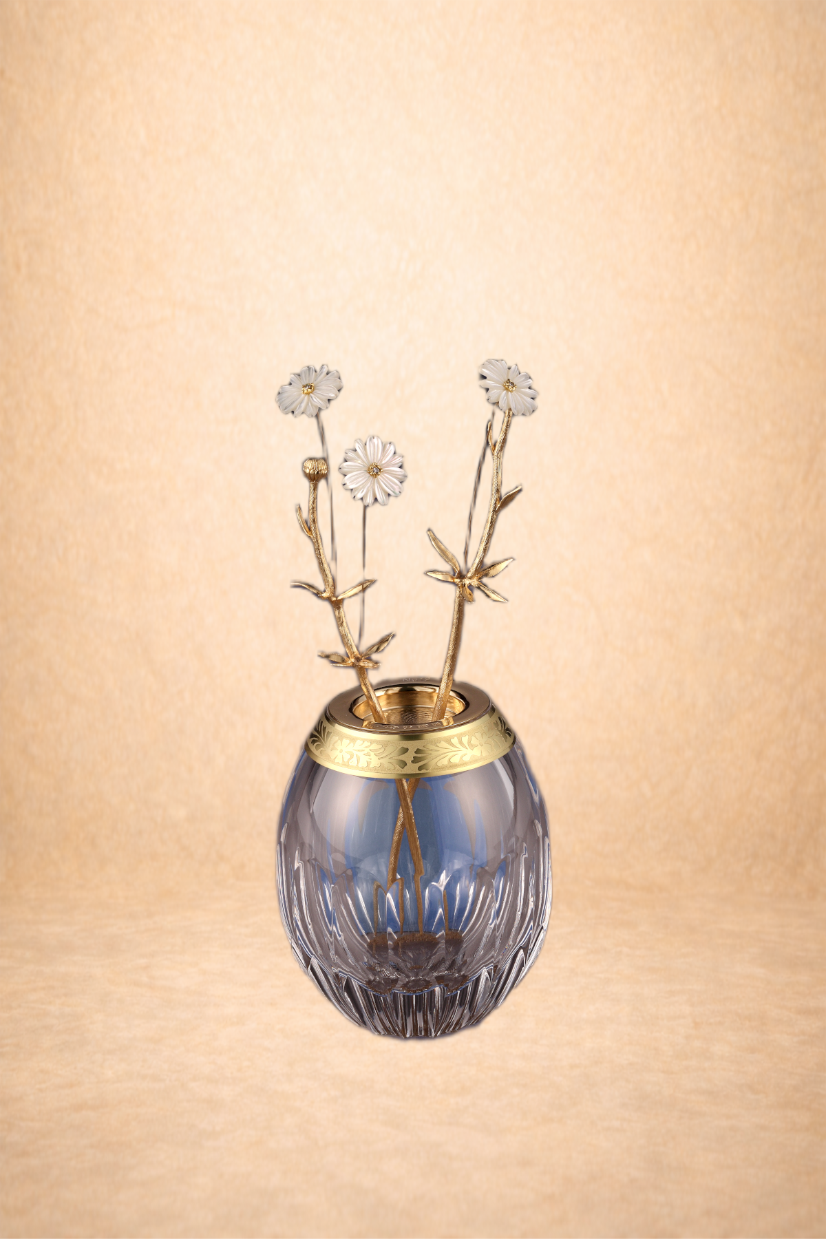 A glass vase with gold accents holding white flower-shaped jewelry or decorations on gold stems against a beige background.