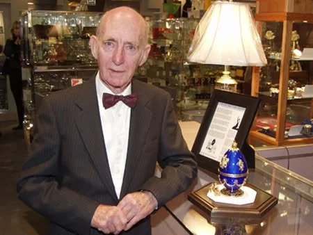 Theo Fabergé standing beside a blue Fabergé egg creation displayed in a glass showcase during an exhibition.