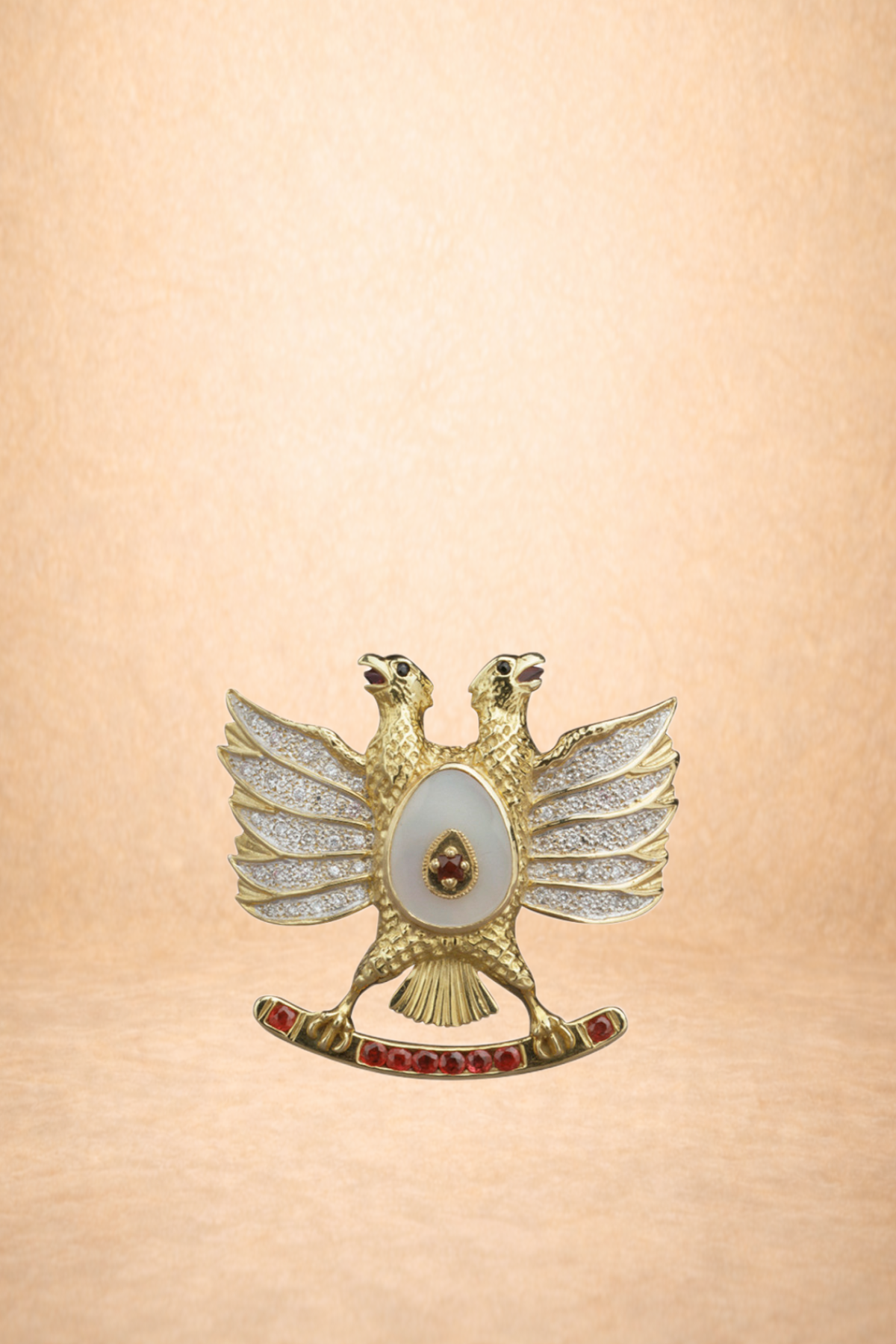 Gold decorative pin shaped like a twin-headed bird with spread wings, jewel accents, and a curved base with red gemstones, set against a soft beige background.