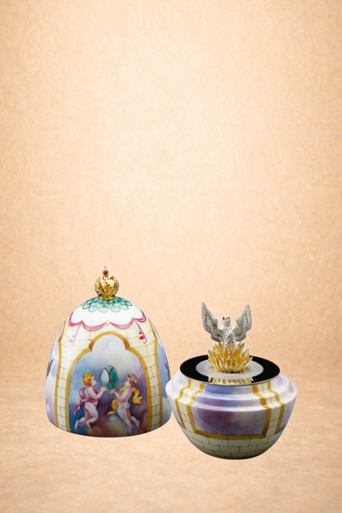 Decorative porcelain vases with painted scenes and gold accents, one with a crown-like ornament and the other with a silver eagle figurine on top, placed against a plain peach background.