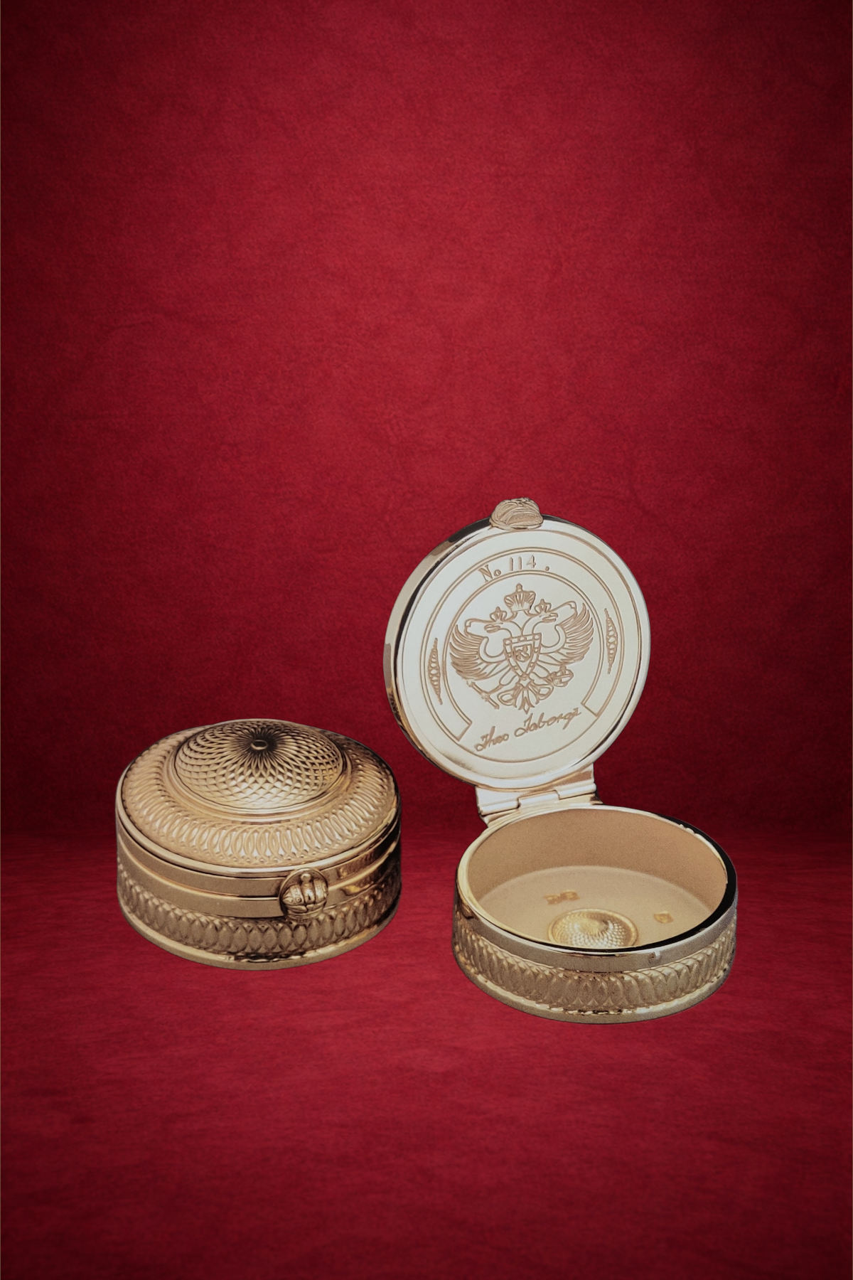 Round, gold-colored, decorative jewelry box with intricate patterns and an embossed lid, placed beside an open matching box with engraved design and initials inside, against a red background.