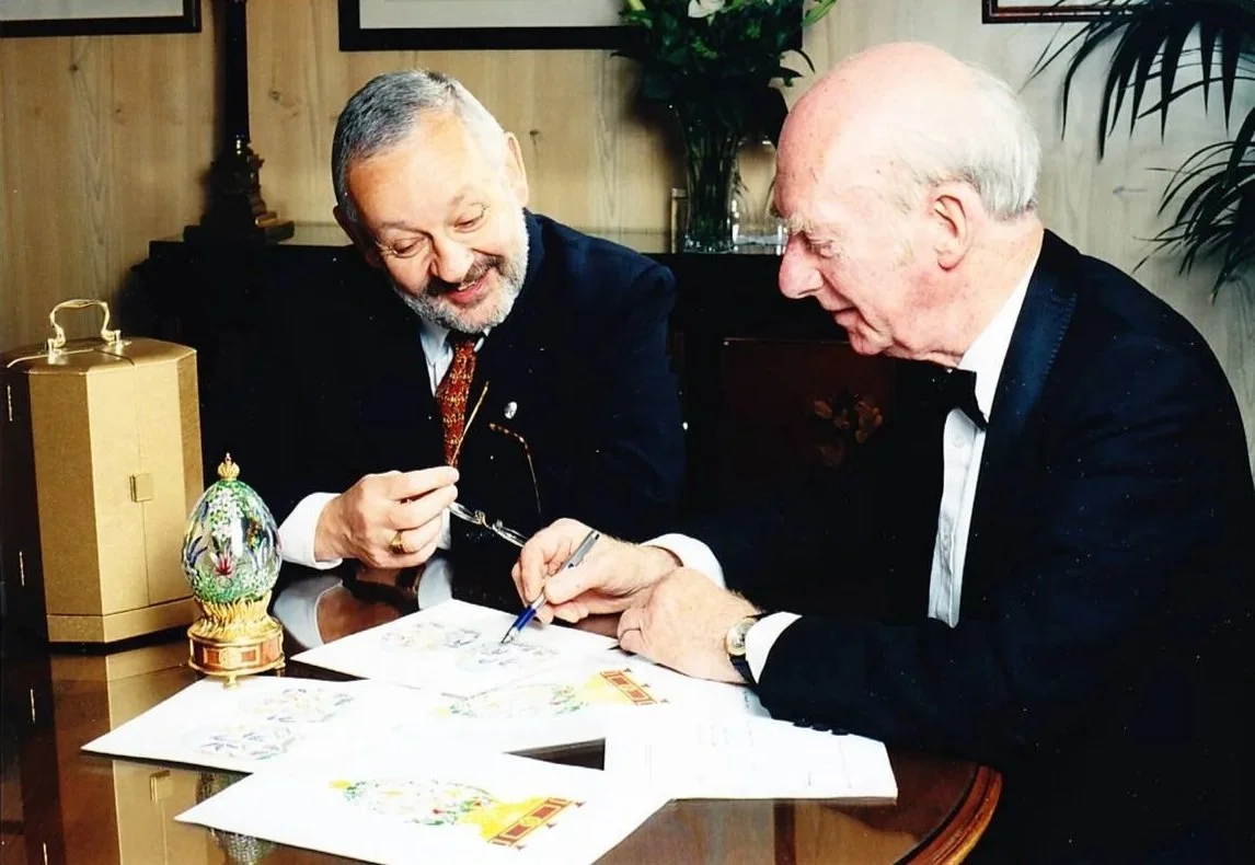 Two men in formal attire are sitting at a table, looking at and discussing colorful artwork or cards. One man is holding glasses and a pin, while the other is writing or drawing. There is a decorative egg-shaped ornament on the table, and a large potted plant and artwork hanging on the wall behind them.