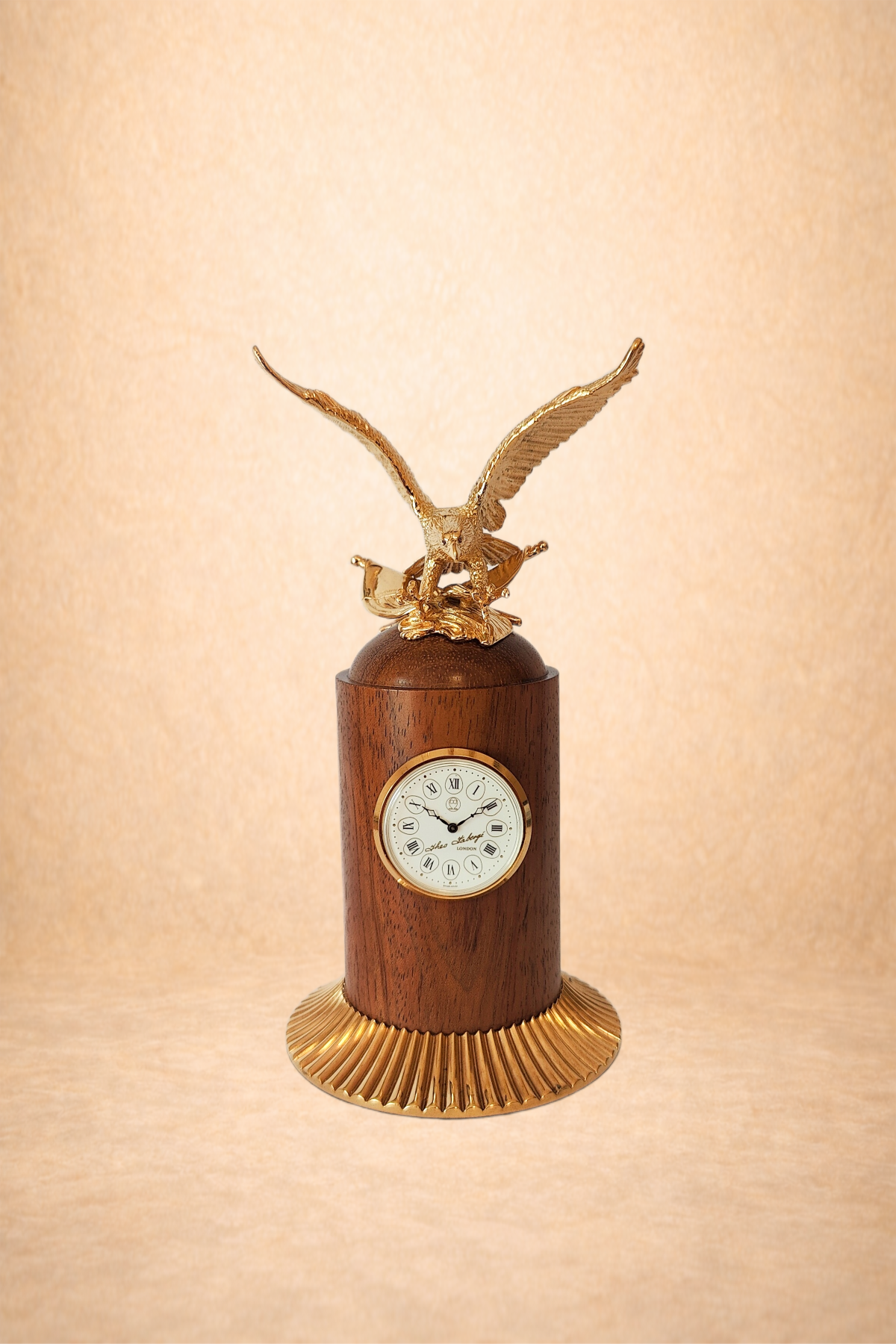 A decorative table clock with a wooden cylindrical base, a gold-colored eagle figurine on top with wings spread, and a round clock face with Roman numerals and black hands.