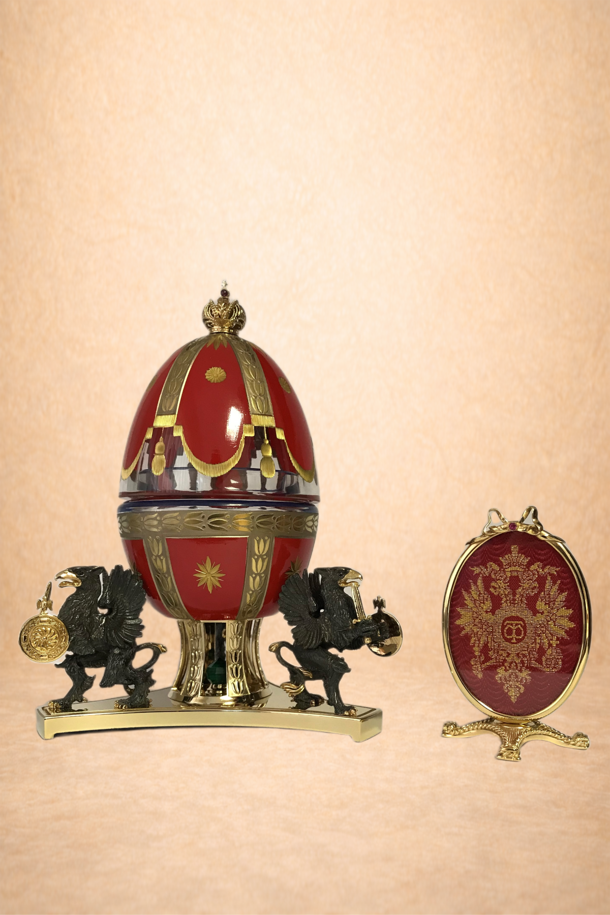 Decorative red and gold Fabergé-style egg with two black lion figurines holding a gold coin and a gold miniature chest on a golden base, and a red and gold embroidered oval frame with a chain on a glossy surface against a beige wall.