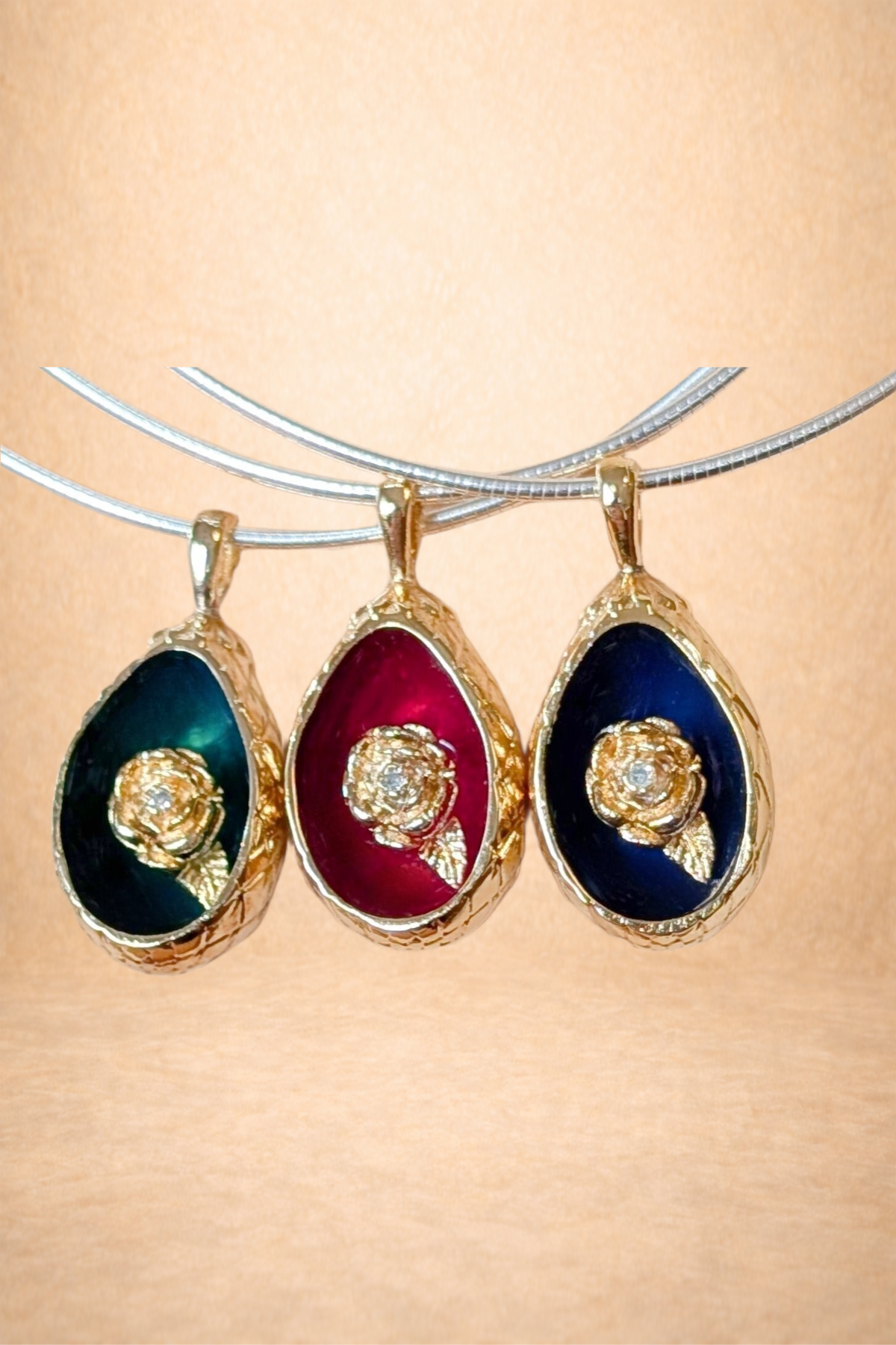Three teardrop-shaped pendants with gold borders and embedded jewelry designs, hanging on a silver curved bar. The pendants feature dark backgrounds in black, red, and blue.