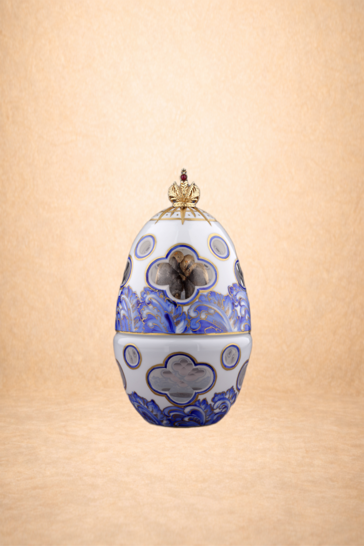 Decorative porcelain egg-shaped container with blue and gold patterns, a crown on top, and a small opening revealing tiny objects inside, set against a beige background.