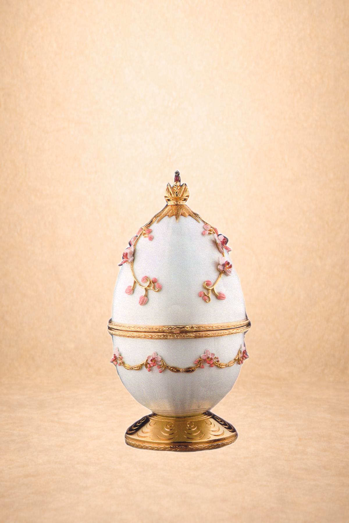 Decorative ornate porcelain egg with gold accents, pink floral details, and a golden crown topper, placed on a gold stand, against a beige background.