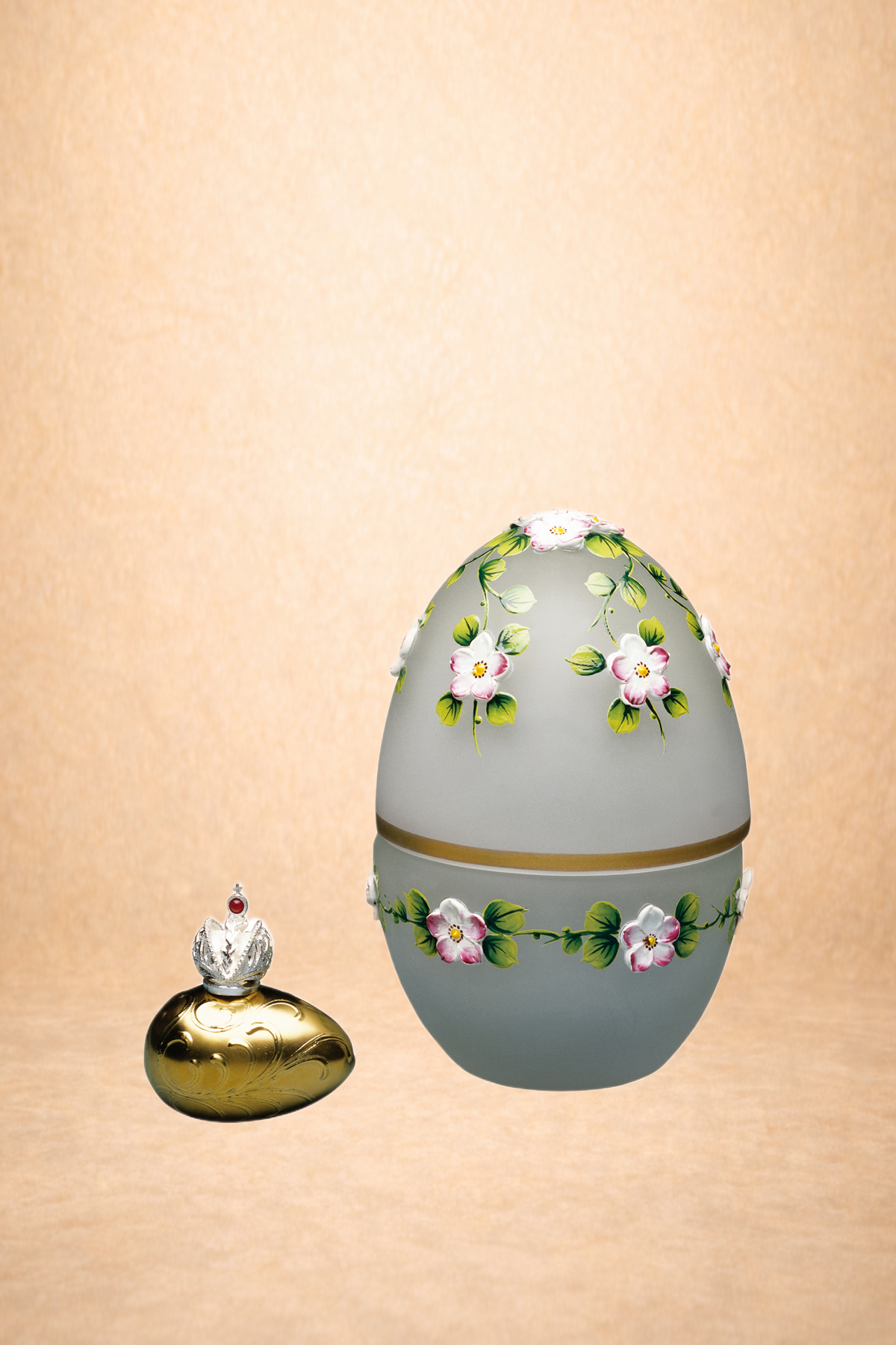 A decorative porcelain egg with pink flowers and green leaves, and a small perfume bottle with a crown-shaped top beside it.