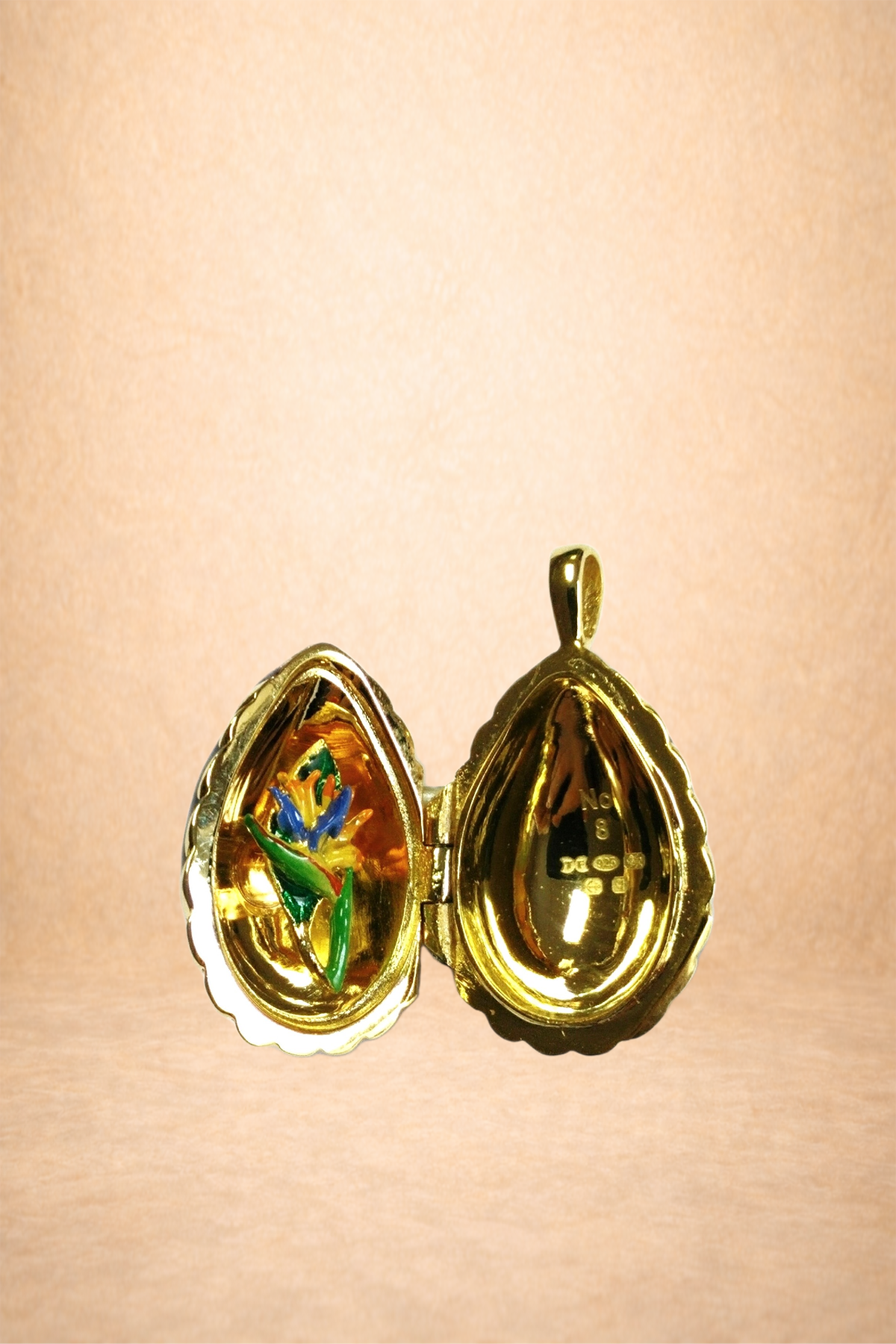 A gold locket open to reveal a colorful enamel flower on one side and engraved text on the other side, hanging against a light pink background.