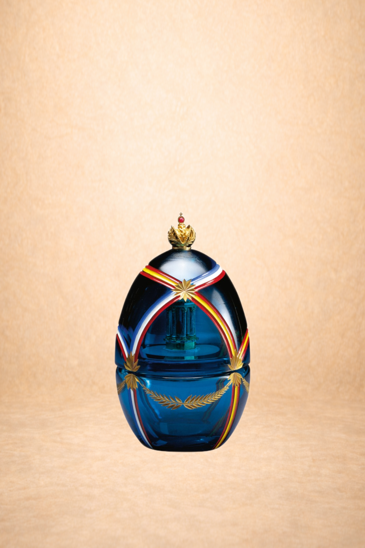A decorative blue glass bottle with gold, red, white, and black ribbon-like accents and a gold crown on top, set against a neutral beige background.