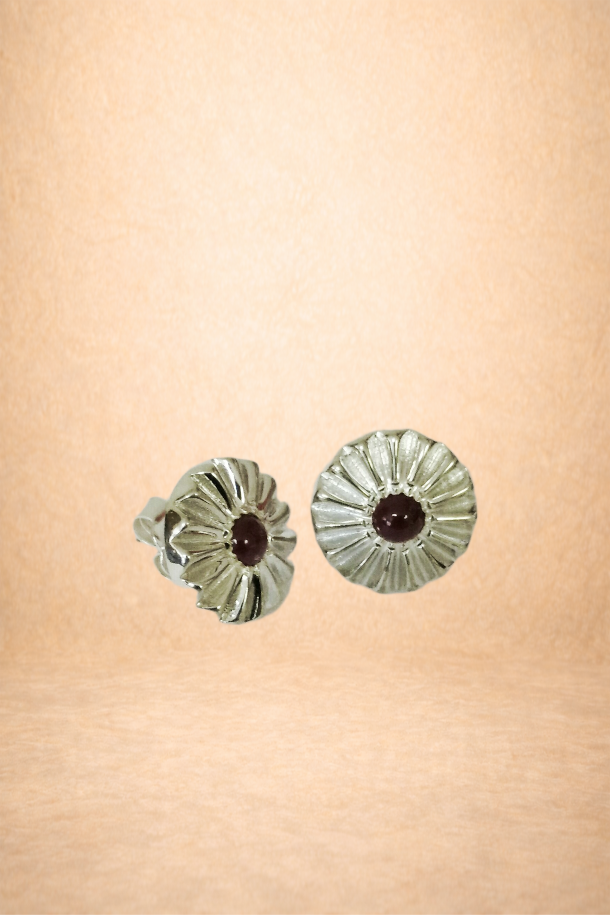 A pair of silver earrings with sunflower design and dark gemstones in the center on a peach-colored background.