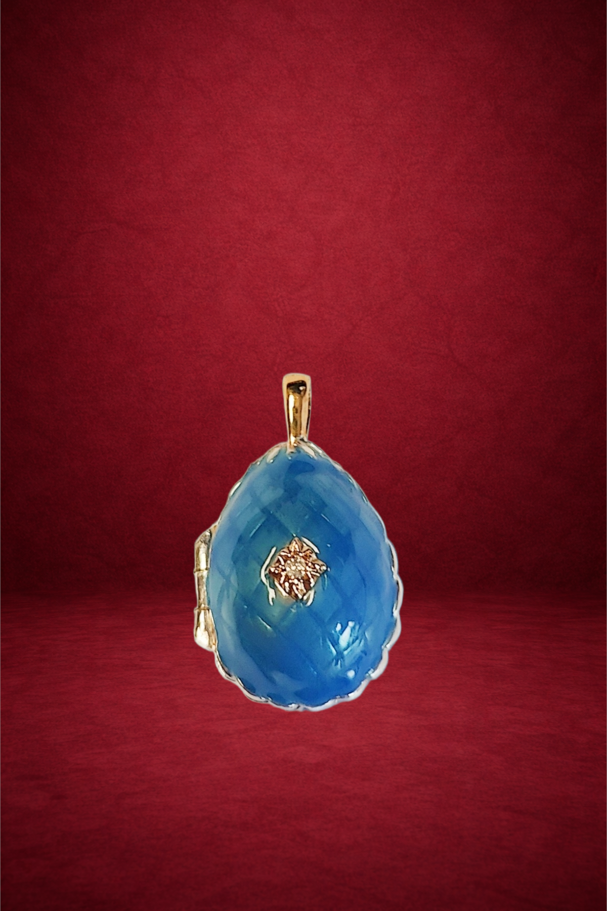 A teardrop-shaped pendant featuring a large blue gemstone with a small diamond set in the center, set in a gold or gold-colored metal frame, displayed against a red background.