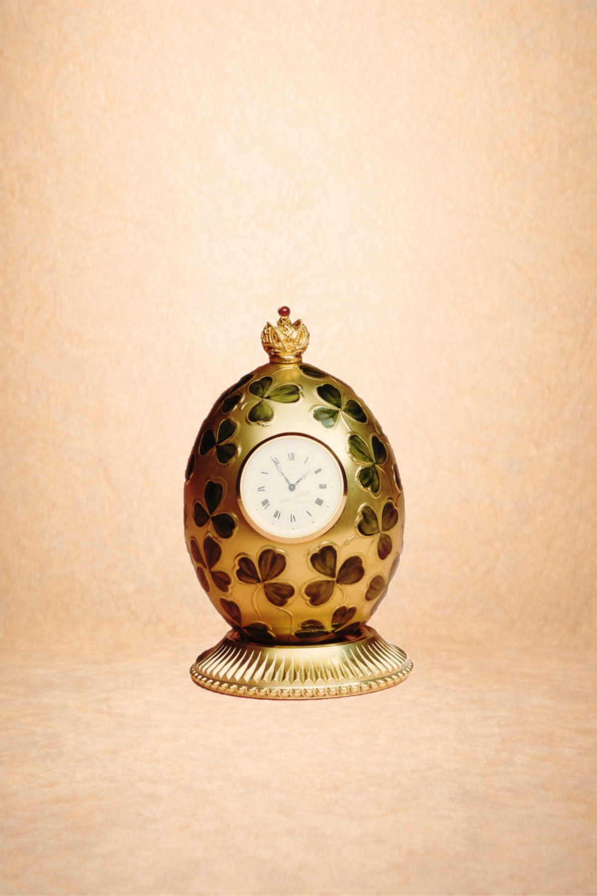 An ornate egg-shaped clock with a white clock face and Roman numerals, decorated with green leaves and topped with a gold crown, on a pedestal.