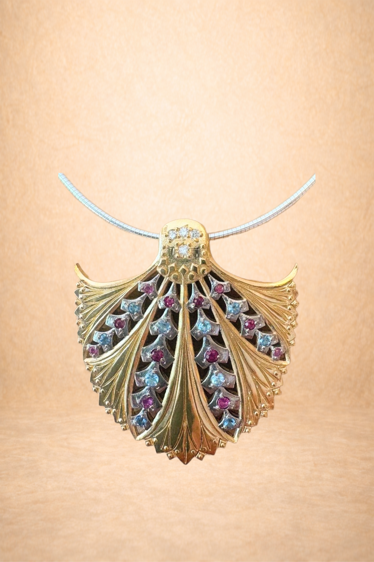 Gold pendant necklace with leaf design and embedded red and clear gemstones, hanging on a silver chain against a beige background.