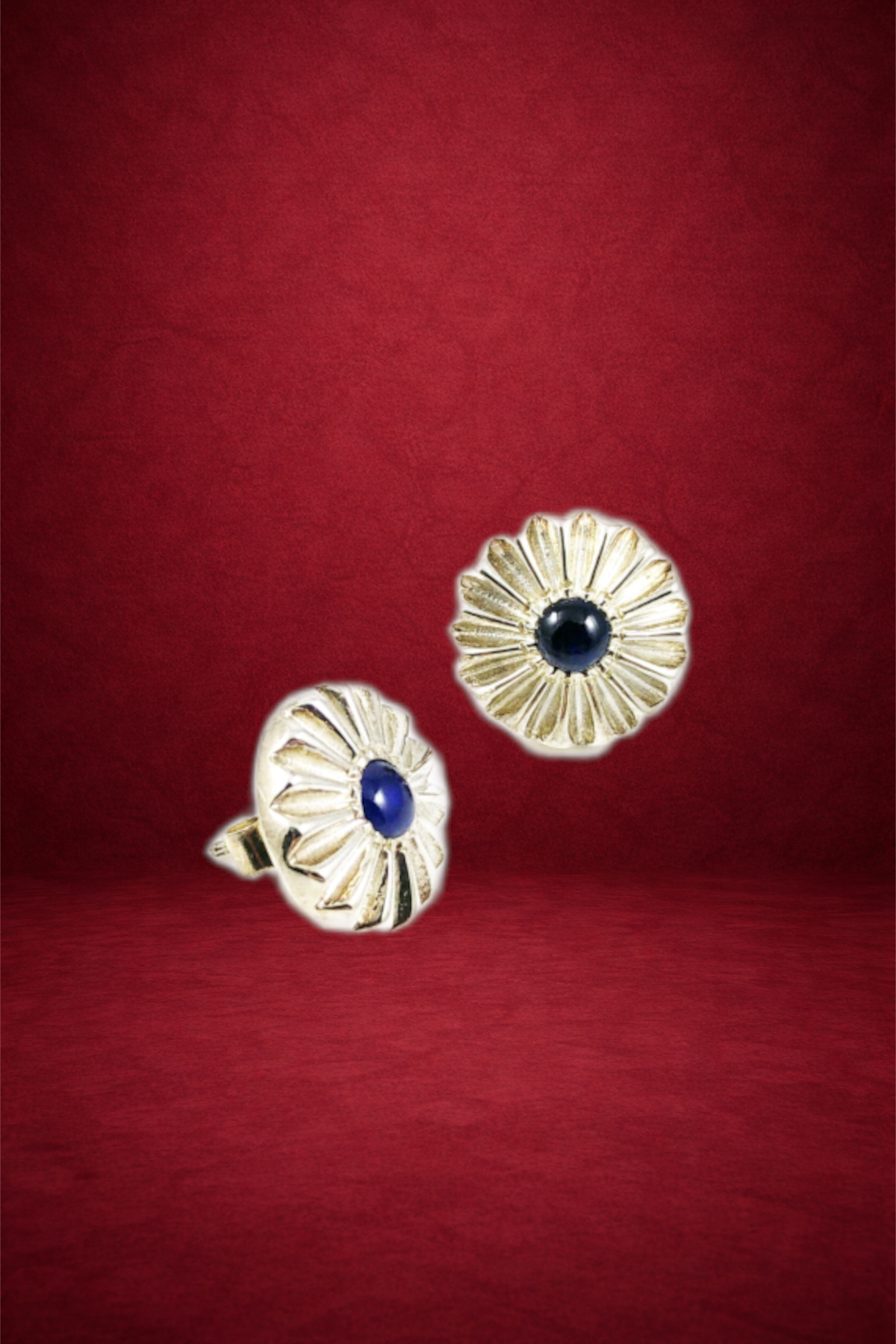 Pair of gold flower-shaped earrings with dark blue stones in the center, placed on a red textured background.