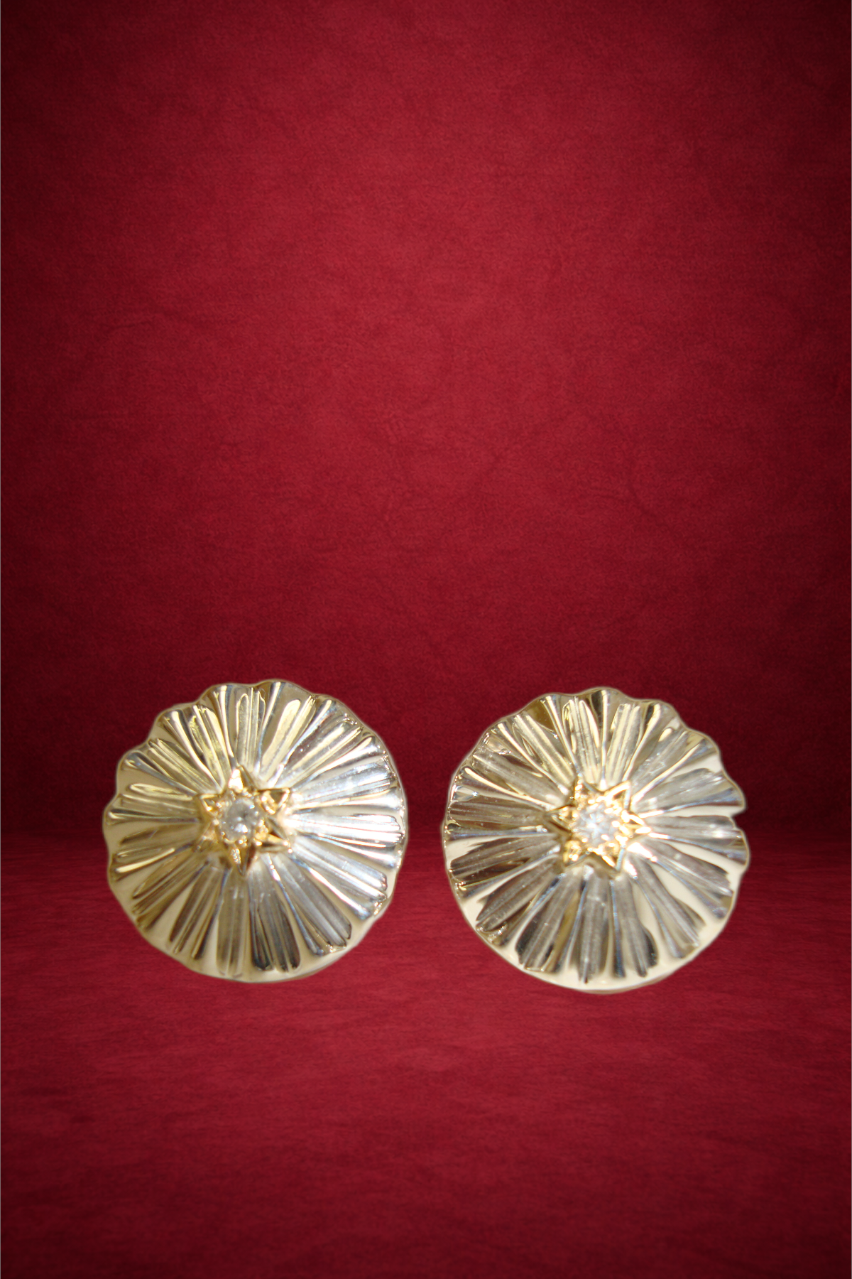 Pair of gold earrings with a flower-like design and a central diamond, placed on a red fabric background.