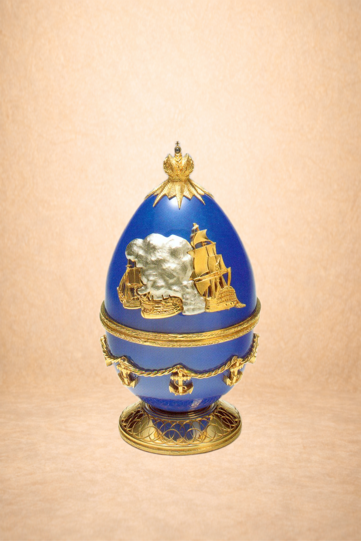 A decorative blue and gold egg with a sailing ship design on its surface, topped with a gold crown and standing on an ornate gold base.