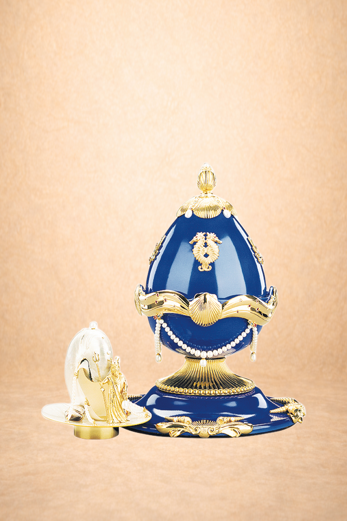 Decorative blue and gold ornate object resembling a Fabergé-style egg with gold embellishments and pearl accents, placed on a matching base, accompanied by a smaller gold and silver decorative insignia or emblem with a swan design, all on a beige background.