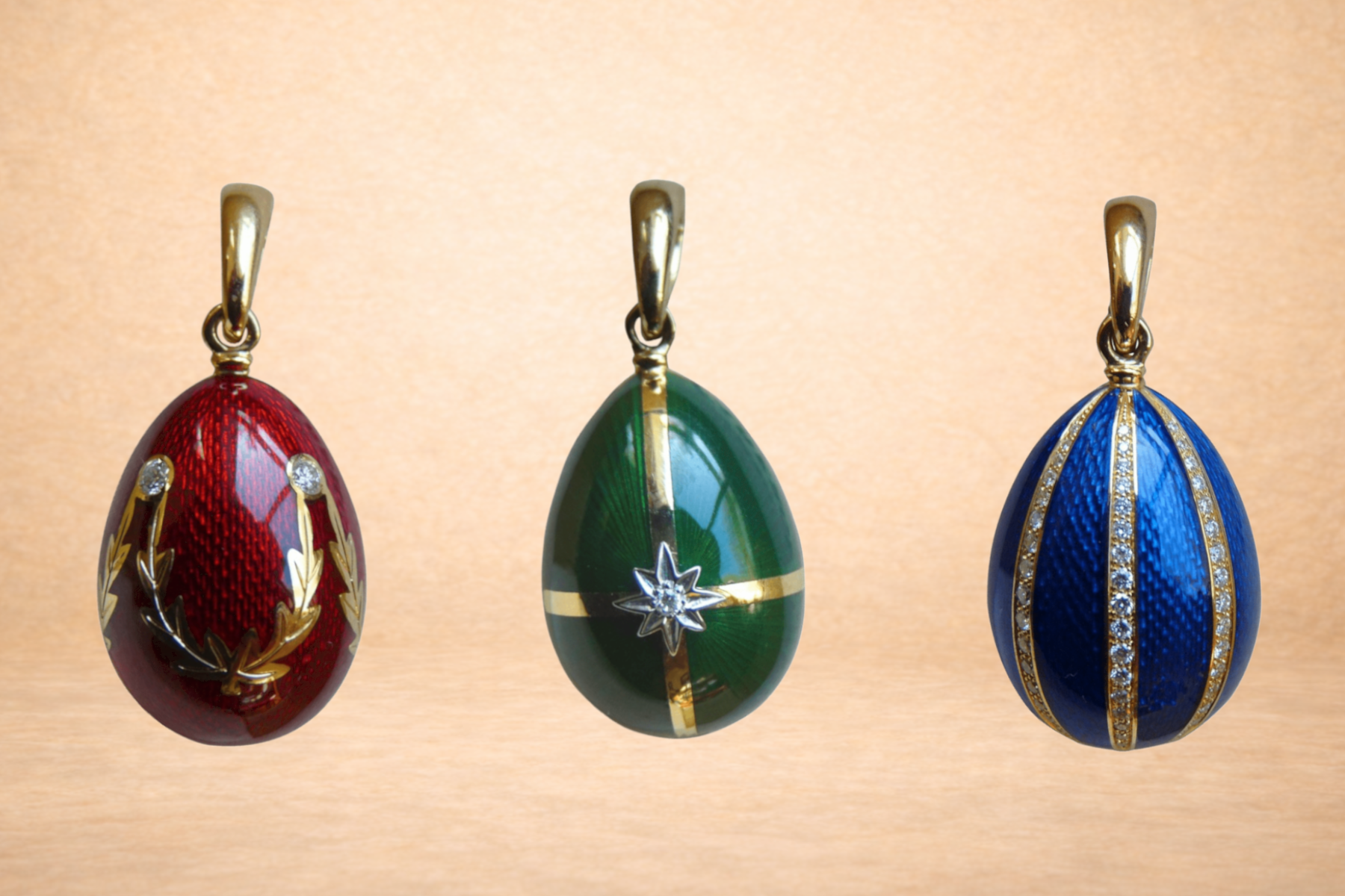 Three decorative egg pendants with gold clasps, red with gold wreath, green with star, and blue with gold stripes, against a beige background.