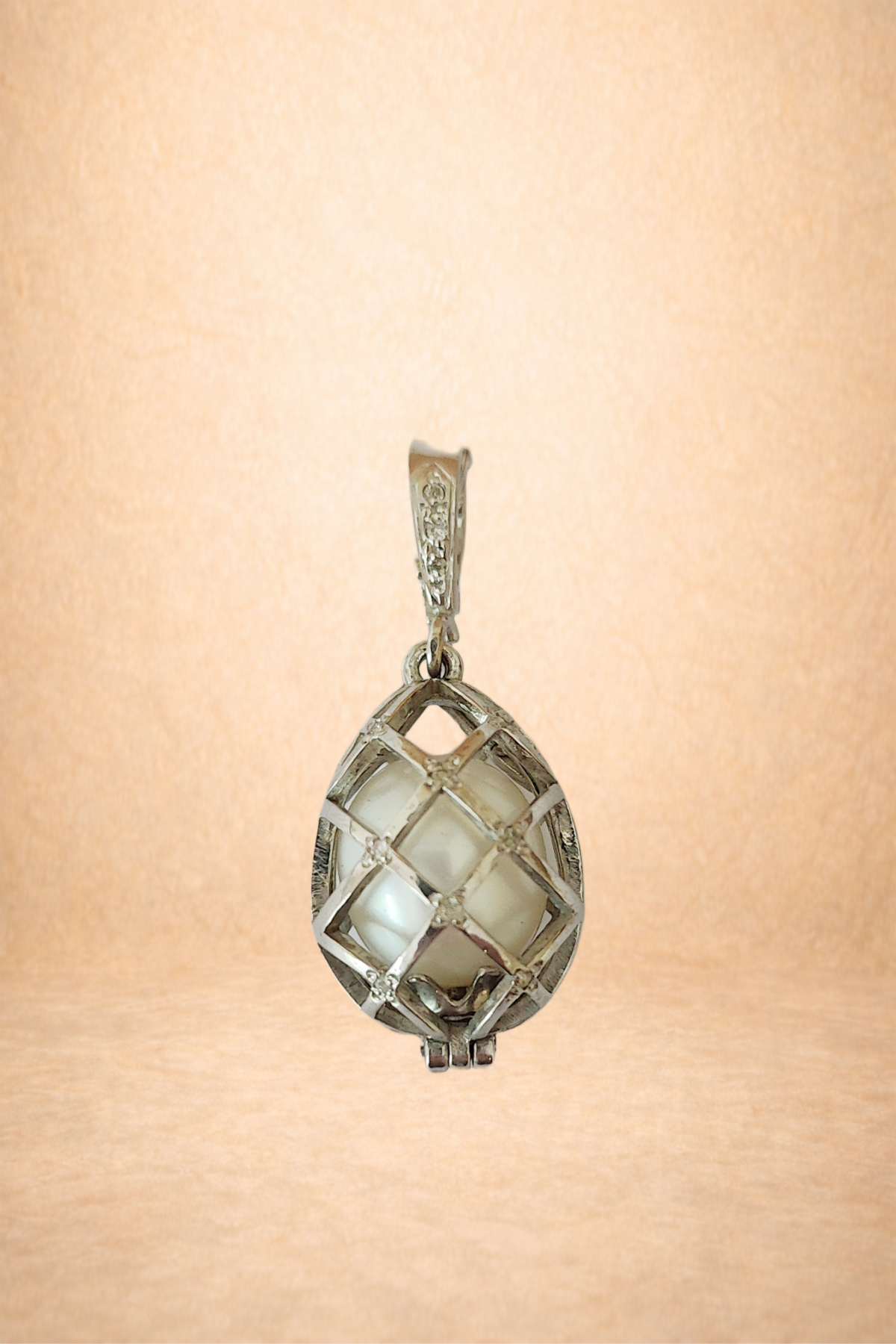 A silver pendant with a square, cage-like design surrounding a round, white, pearl-like stone, hanging against a soft peach-colored background.