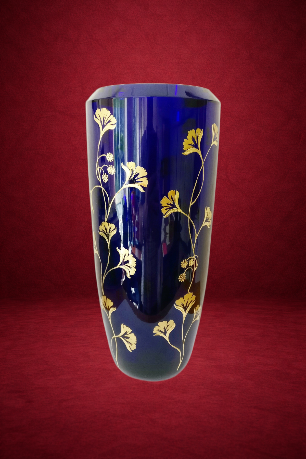 A tall, cylindrical cobalt blue glass vase decorated with gold floral patterns against a deep red background.