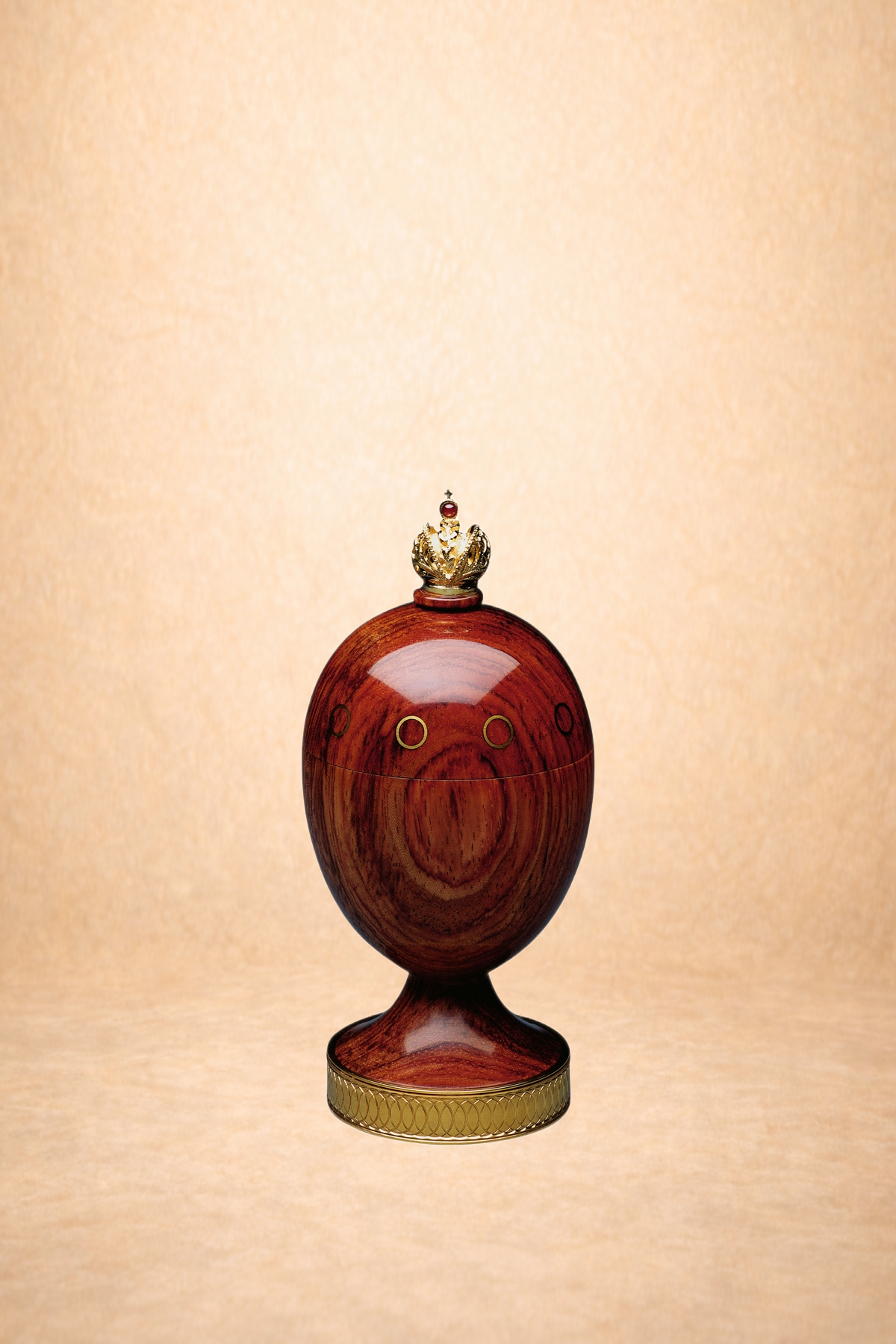 A wooden egg-shaped object with small circular openings and a gold crown on top, resting on a decorative gold stand against a light background.