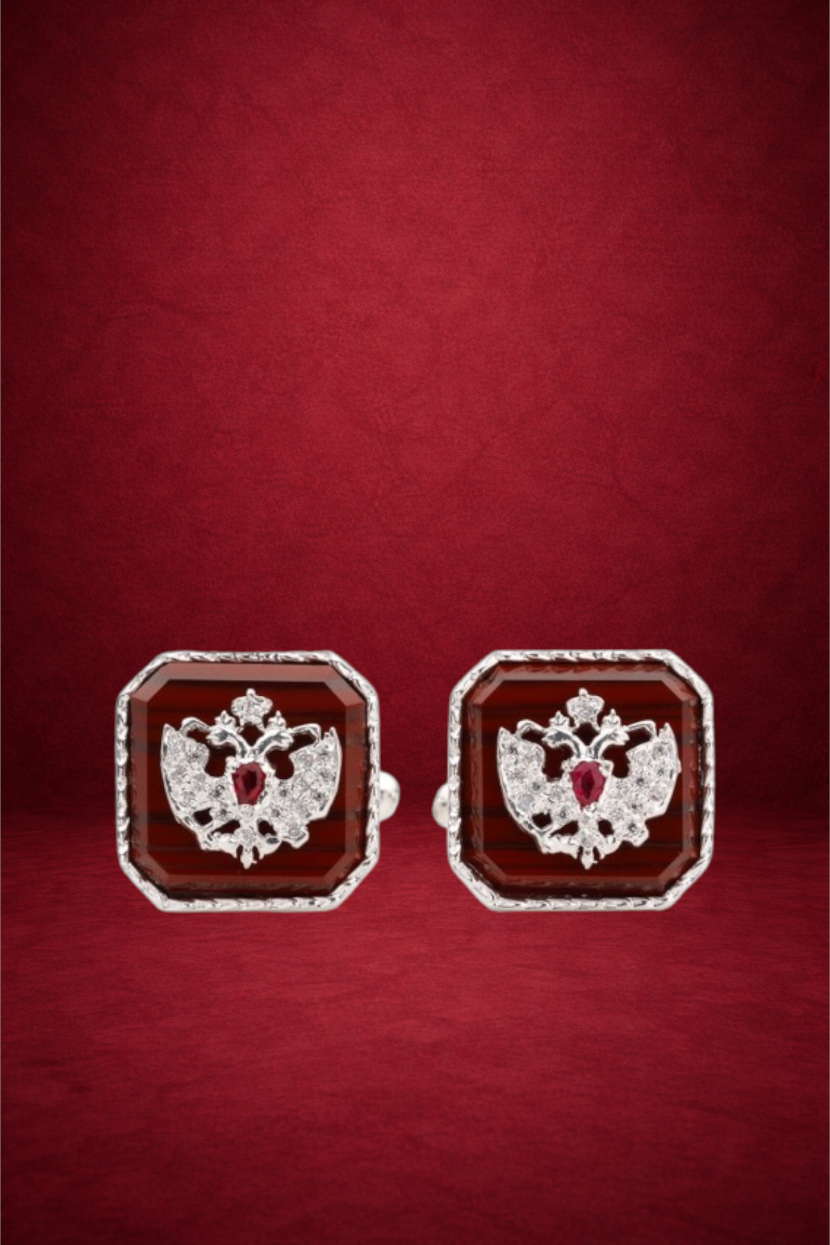 Pair of silver earrings with red gemstone and intricate double-headed eagle design, set against a red background.