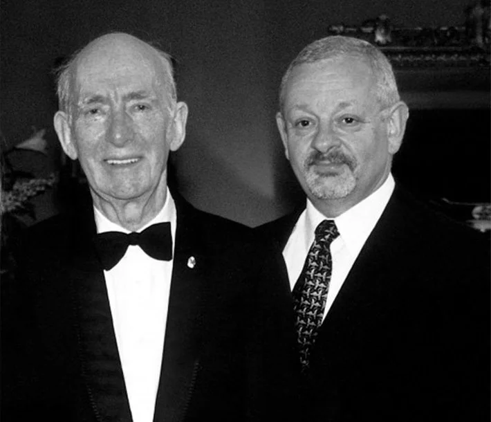 Two middle-aged men in formal tuxedos posing together at a formal event.