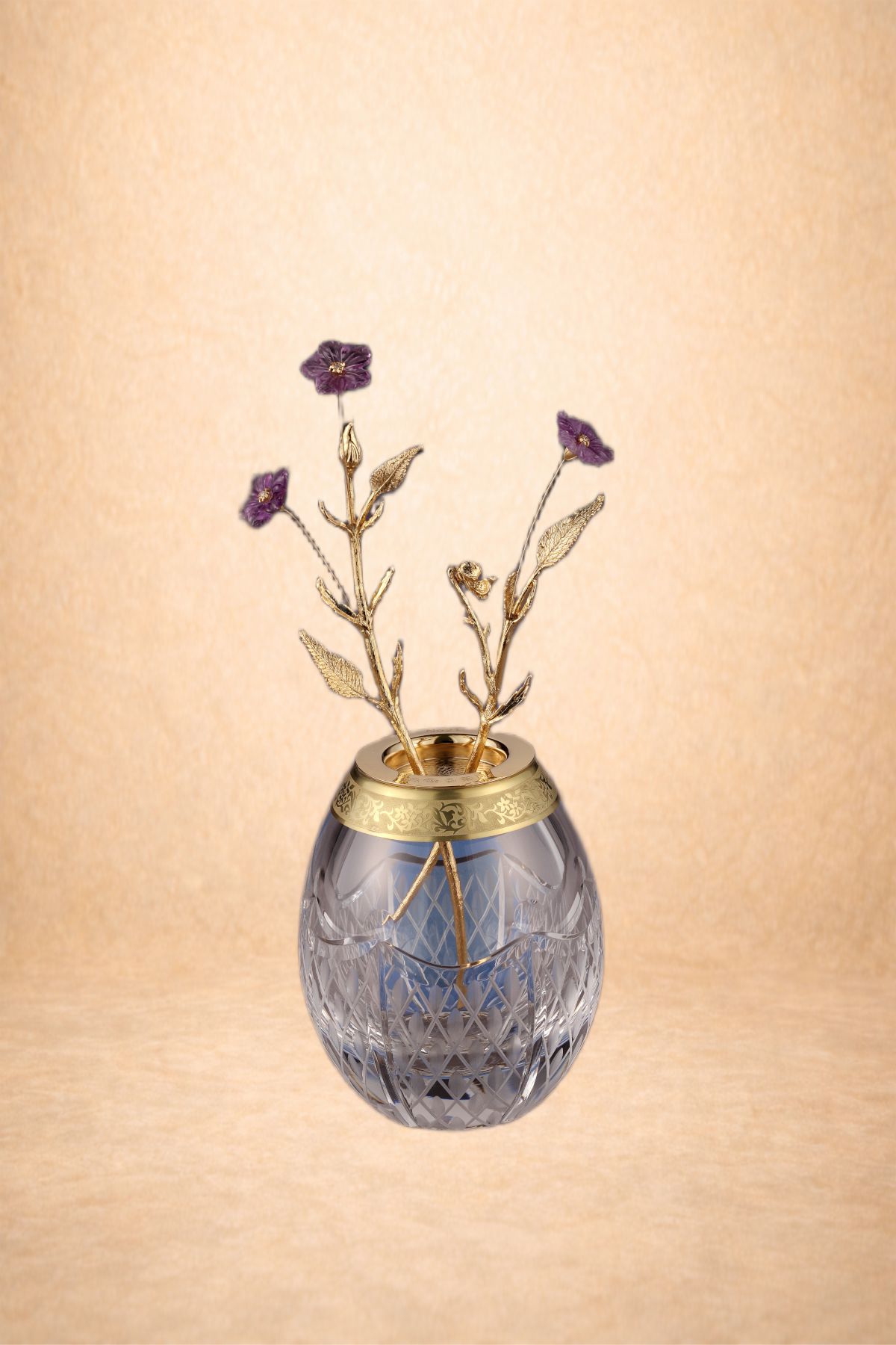 A decorative glass vase with gold accents holding purple embroidered flowers with gold stems and leaves.