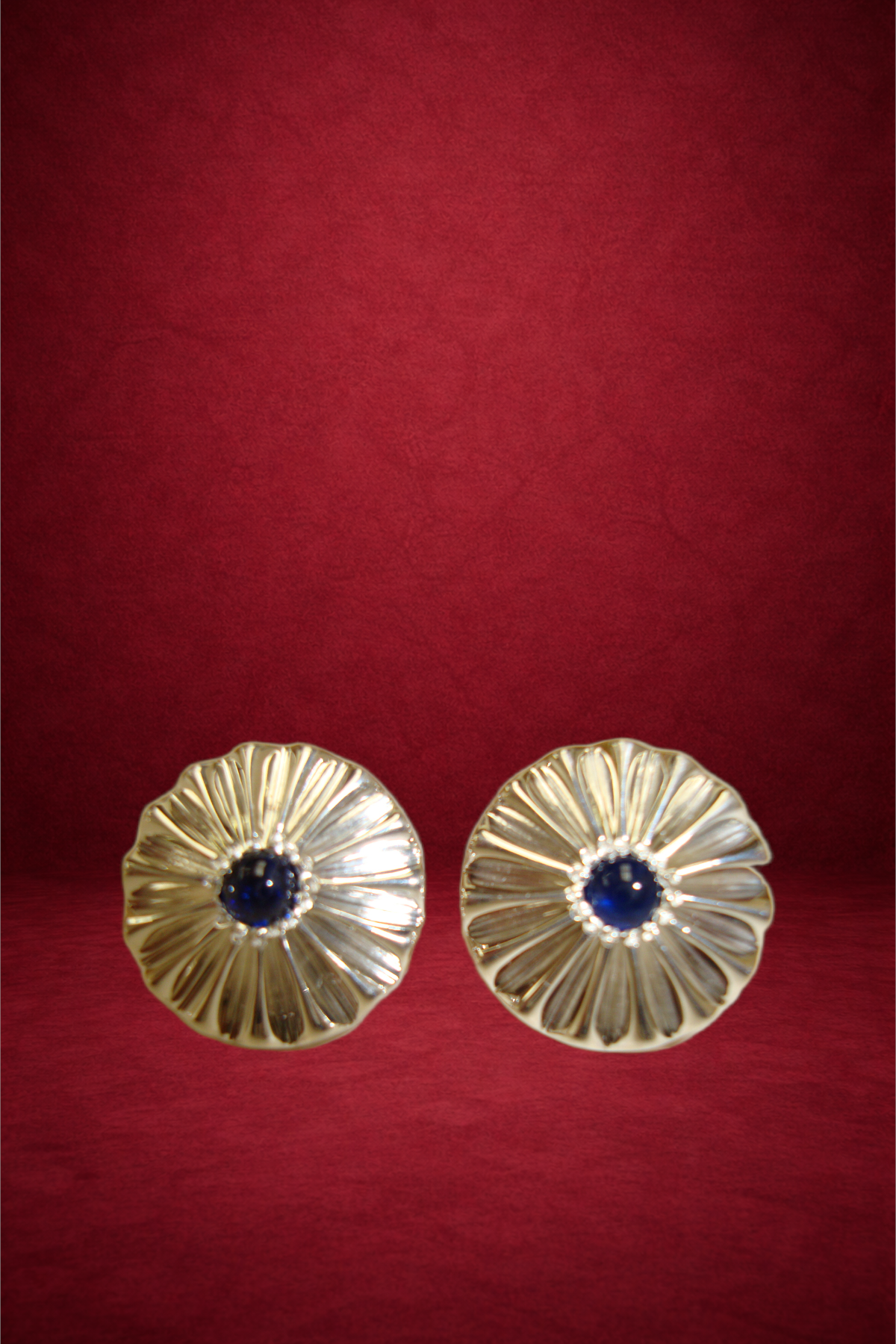 Pair of gold earrings with a central blue gemstone, showcasing a ridged, floral design, against a red background.