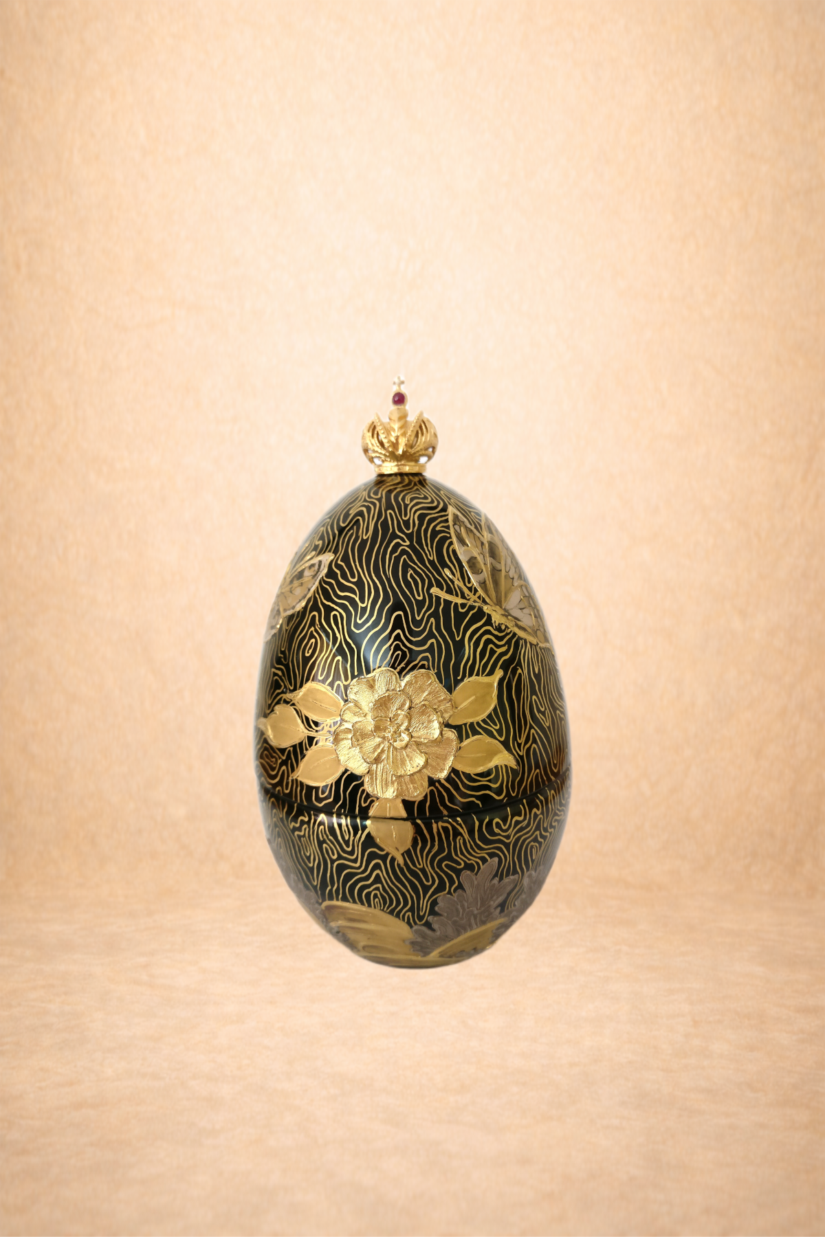 A decorative black and gold egg with floral designs and a gold crown on top, reflected on a shiny surface against a neutral background.
