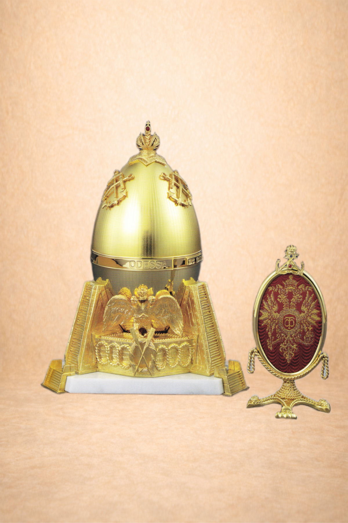A gold decorative egg-shaped object with military insignia and a scepter on top, placed on a white base, beside a small ornate gold frame with red fabric and a gold double-headed eagle emblem.