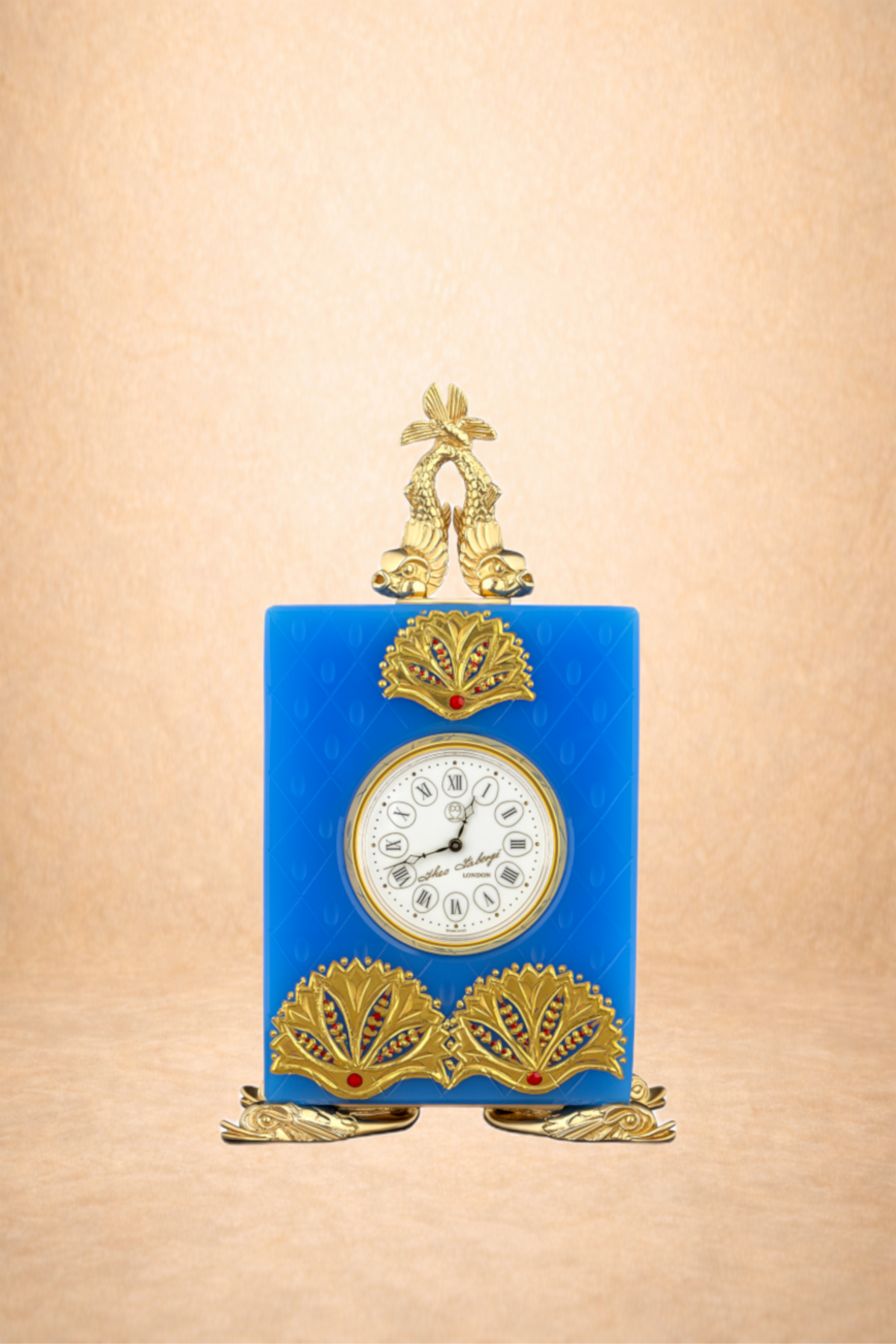 A decorative clock with a blue rectangular body, gold ornamental accents, and a white clock face with Roman numerals. It has a pair of gold fish at the top and gold fan-like designs at the bottom, set against a beige background.