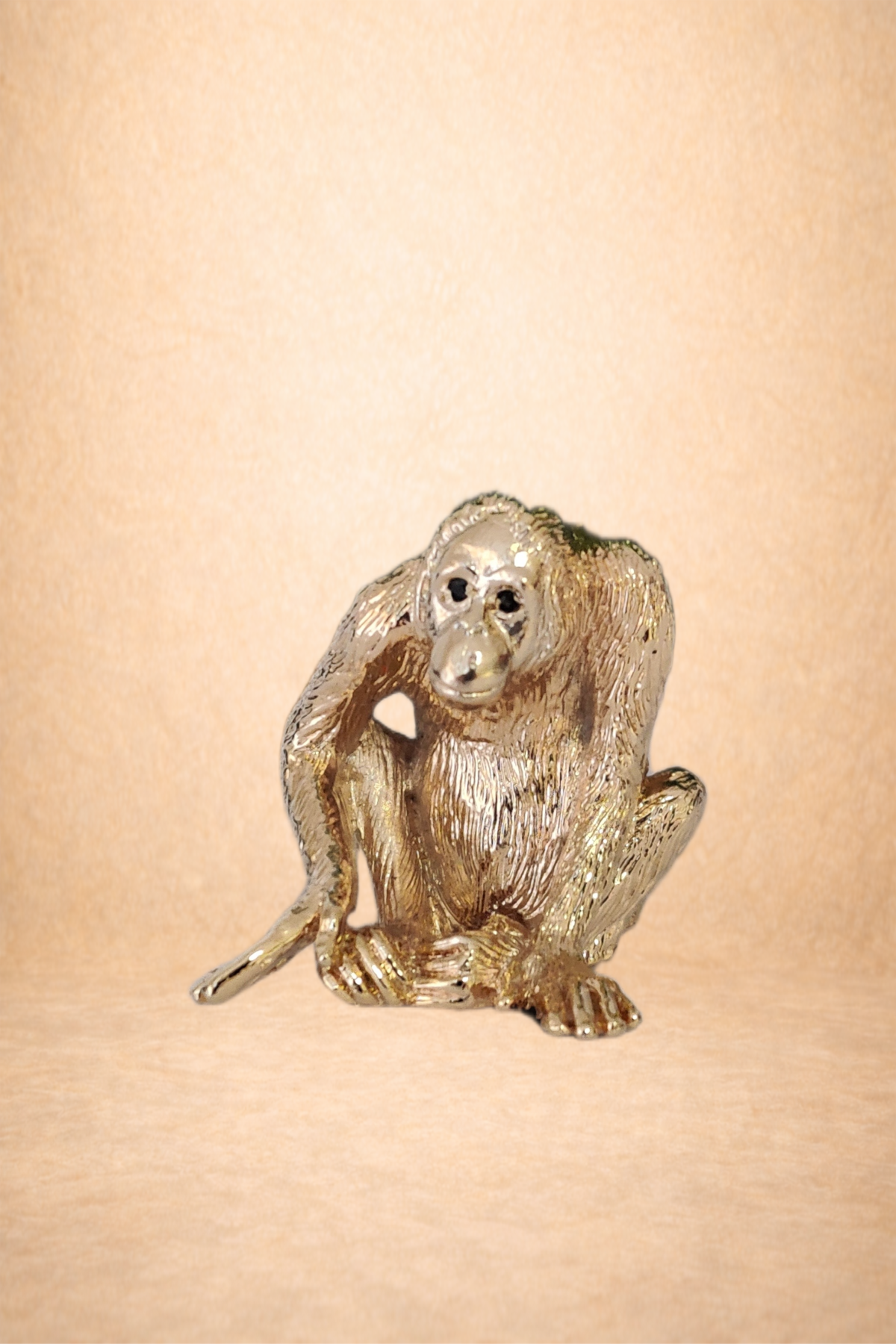 A gold-colored metallic figurine of a monkey, sitting with its body turned and head facing forward, on a plain beige background.