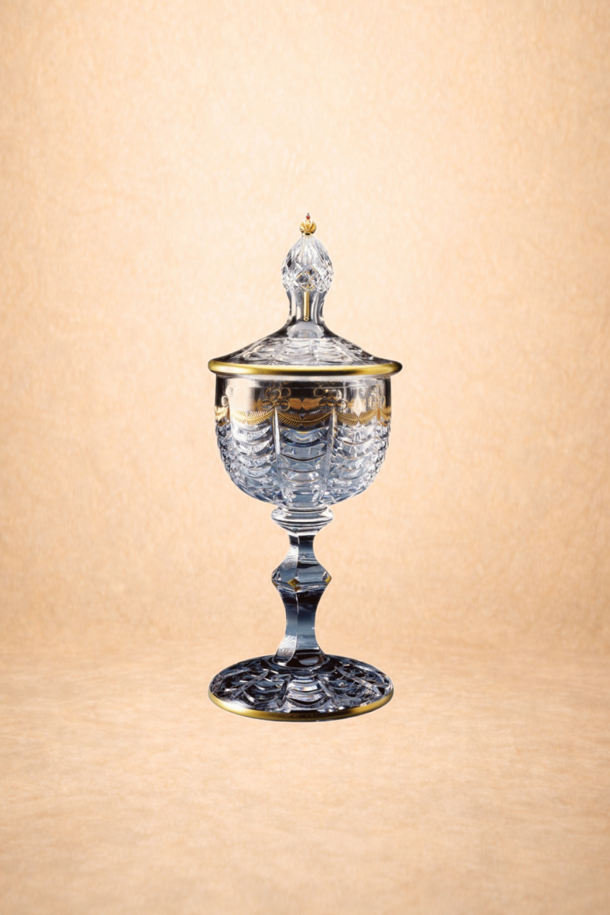 A decorative glass container with a lid featuring gold accents and a pointed finial. The container is positioned on a tall, faceted glass stem with a wide, patterned base, set against a light, textured background.