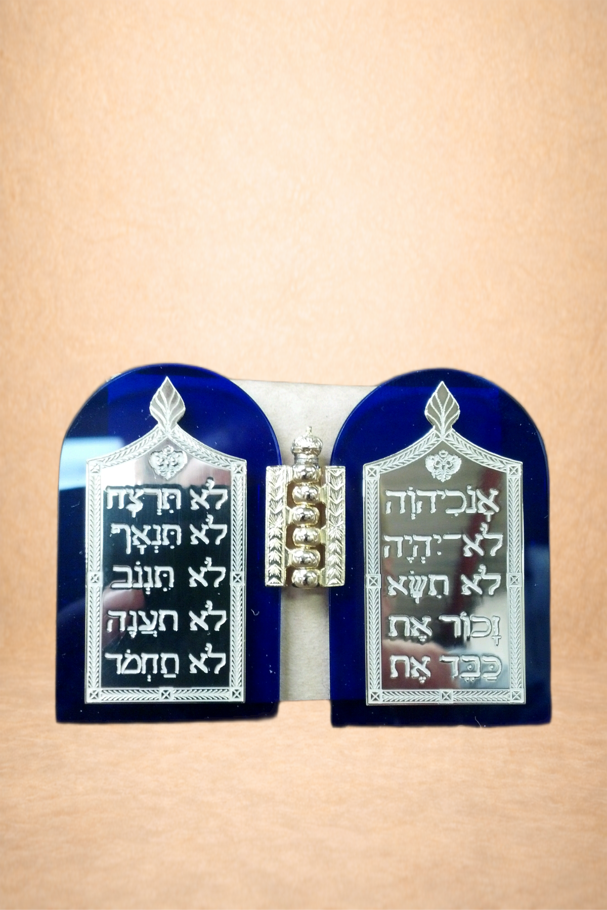 A small decorative item resembling two blue tablets with silver accents, featuring Hebrew inscriptions, separated by a vertical row of four silver balls, on a brown surface.