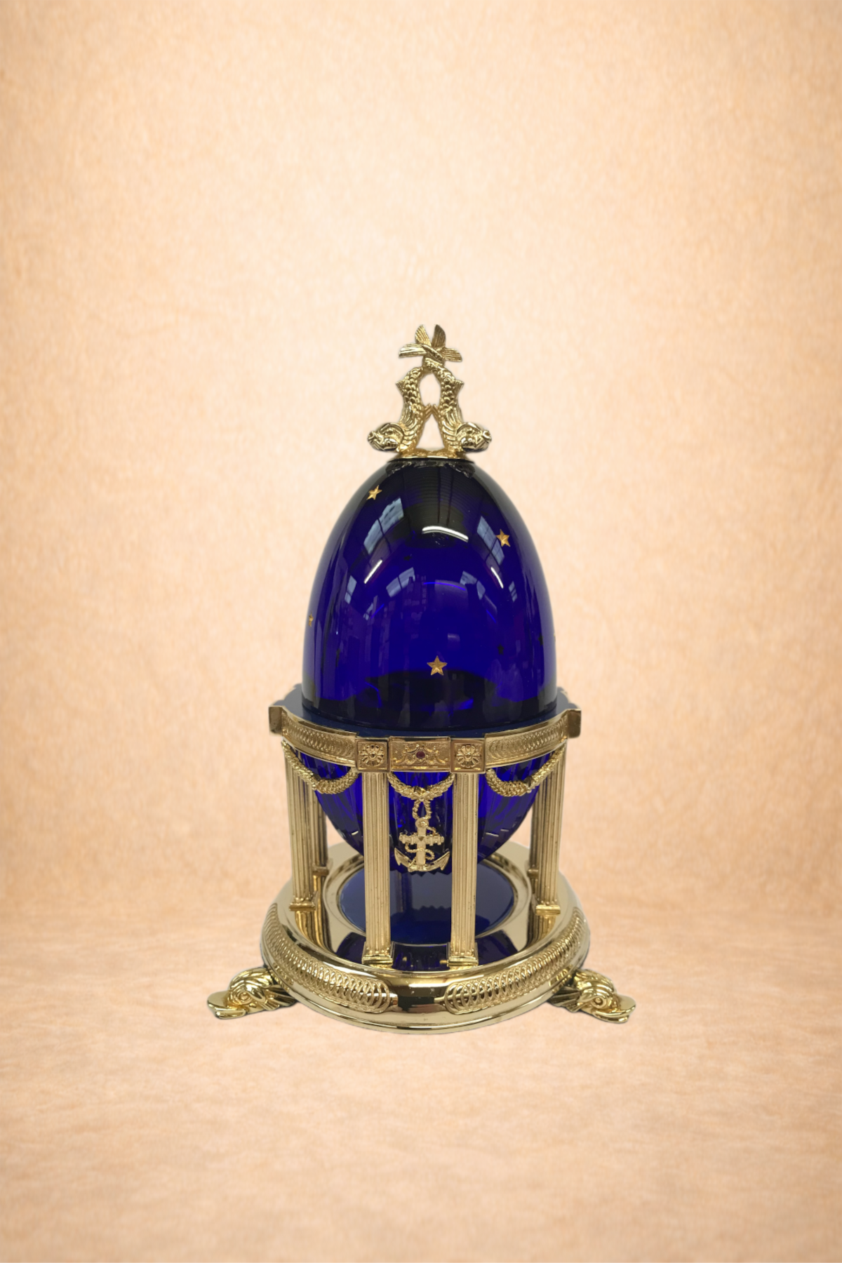 A blue glass egg with gold stars, set on an elaborate gold and silver stand with small columns and a nautical anchor pendant.