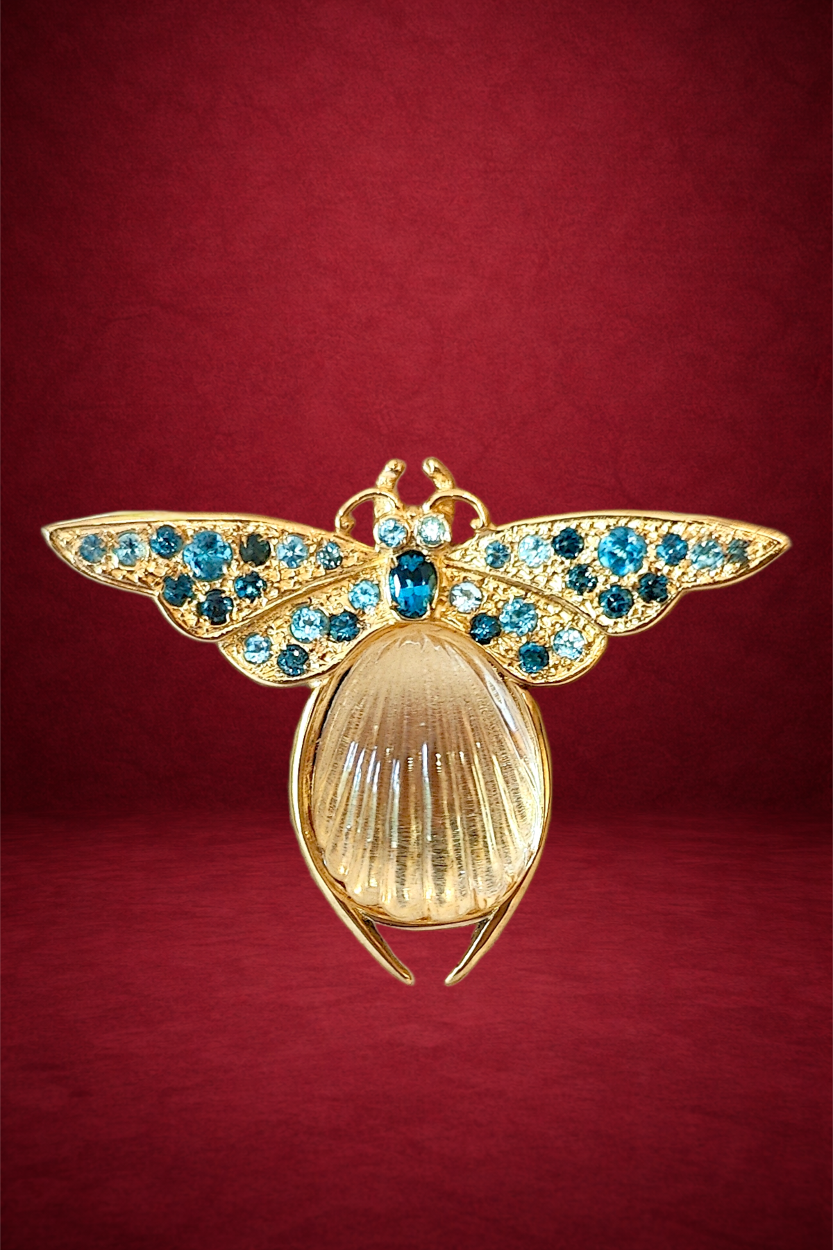 Gold and blue jeweled bee brooch with a shell abdomen on a red background.