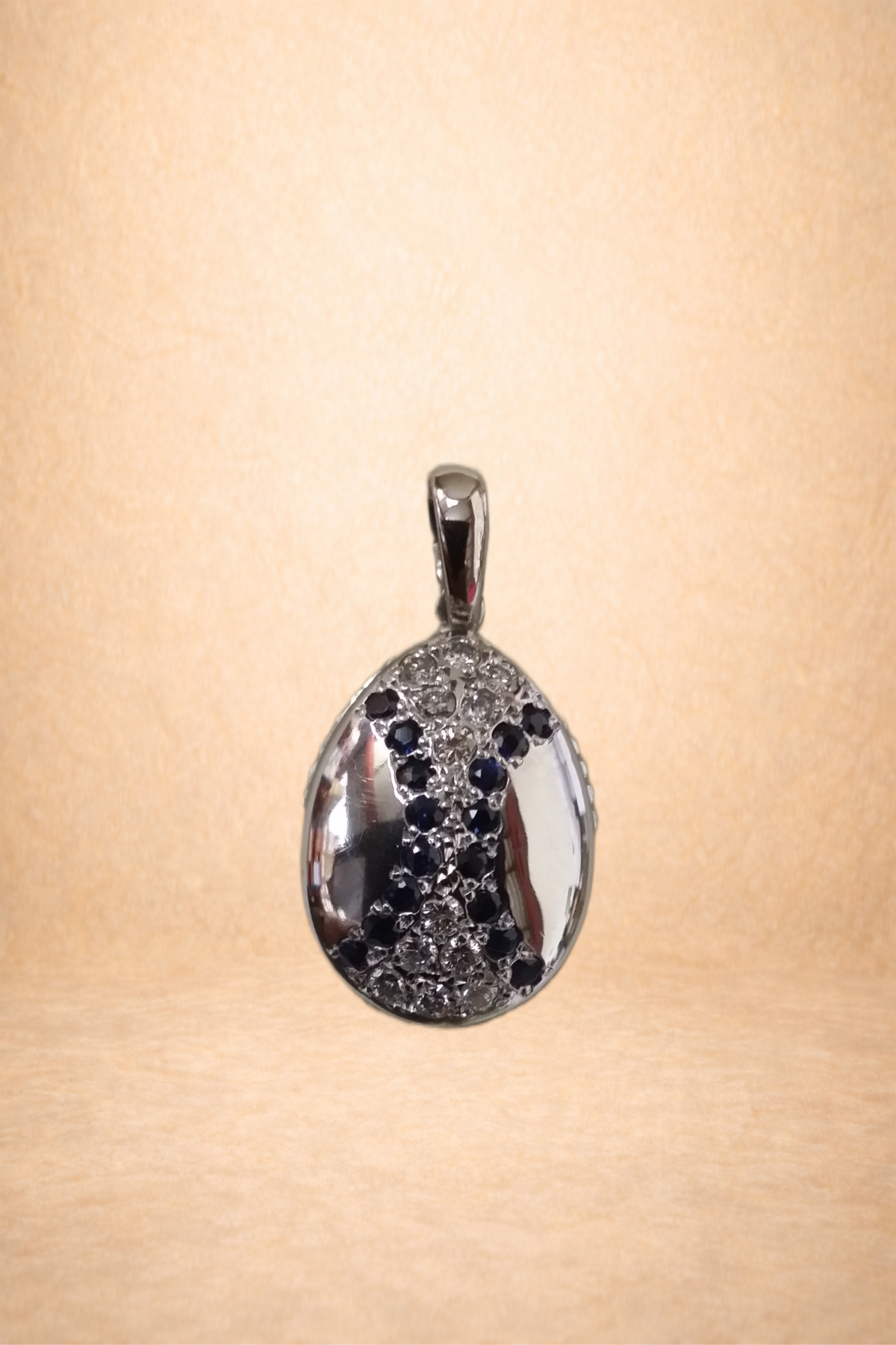 Silver pendant with black and clear gemstones, featuring the letter 'X' on the front, hanging against a beige background.