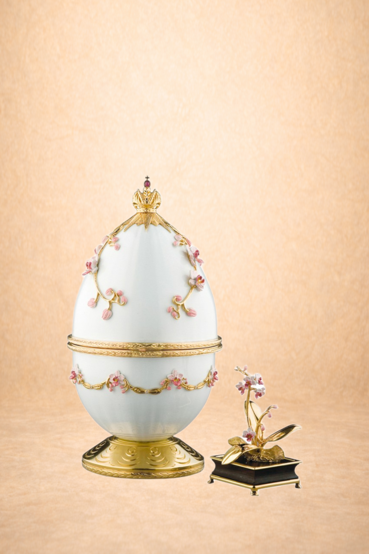 Elegant decorative egg-shaped container with gold accents and pink floral embellishments, topped with a gold crown, accompanied by a small gold and black tray with a gold floral sculpture.