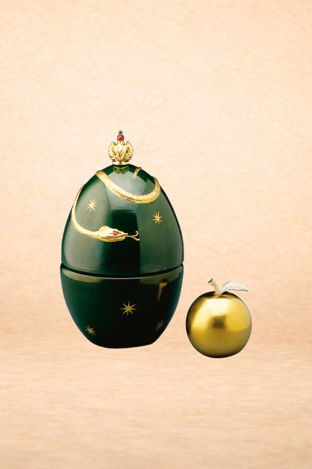 A decorative container shaped like an egg with a dark green color and gold star patterns, topped with a gold crown and jewelry. Next to it is a small, shiny gold apple with a silver leaf.