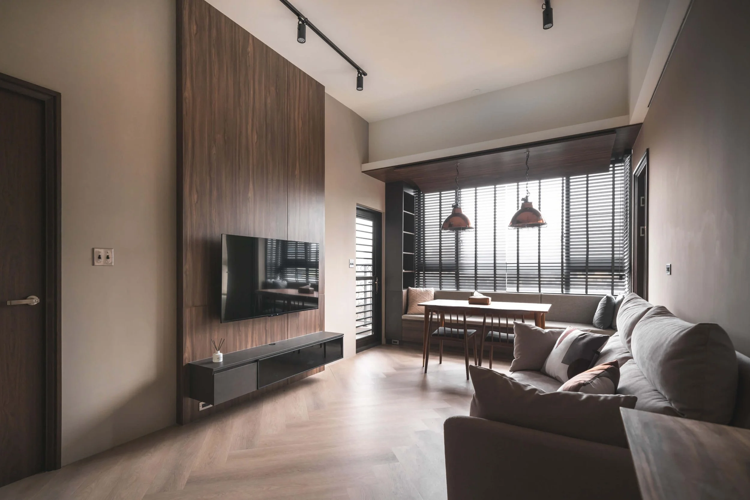 台中 Until 室內設計 P House 客廳，胡桃木紋電視牆與灰色沙發，軌道燈照明，窗邊百葉窗下為餐桌與吊燈