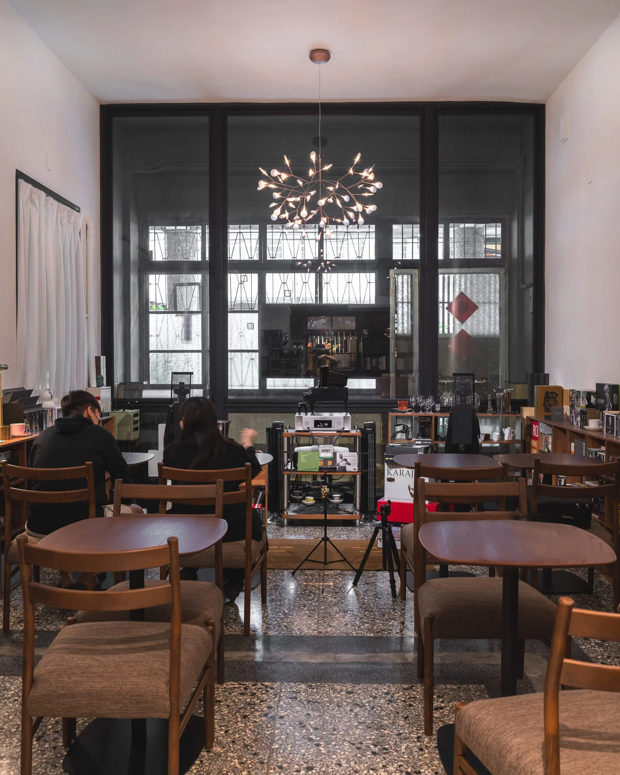 台中 Until 室內設計 Feng Cafe 奉咖啡挑高空間，深色格窗前懸掛枝狀吊燈，木色桌椅排列於磨石子地坪上