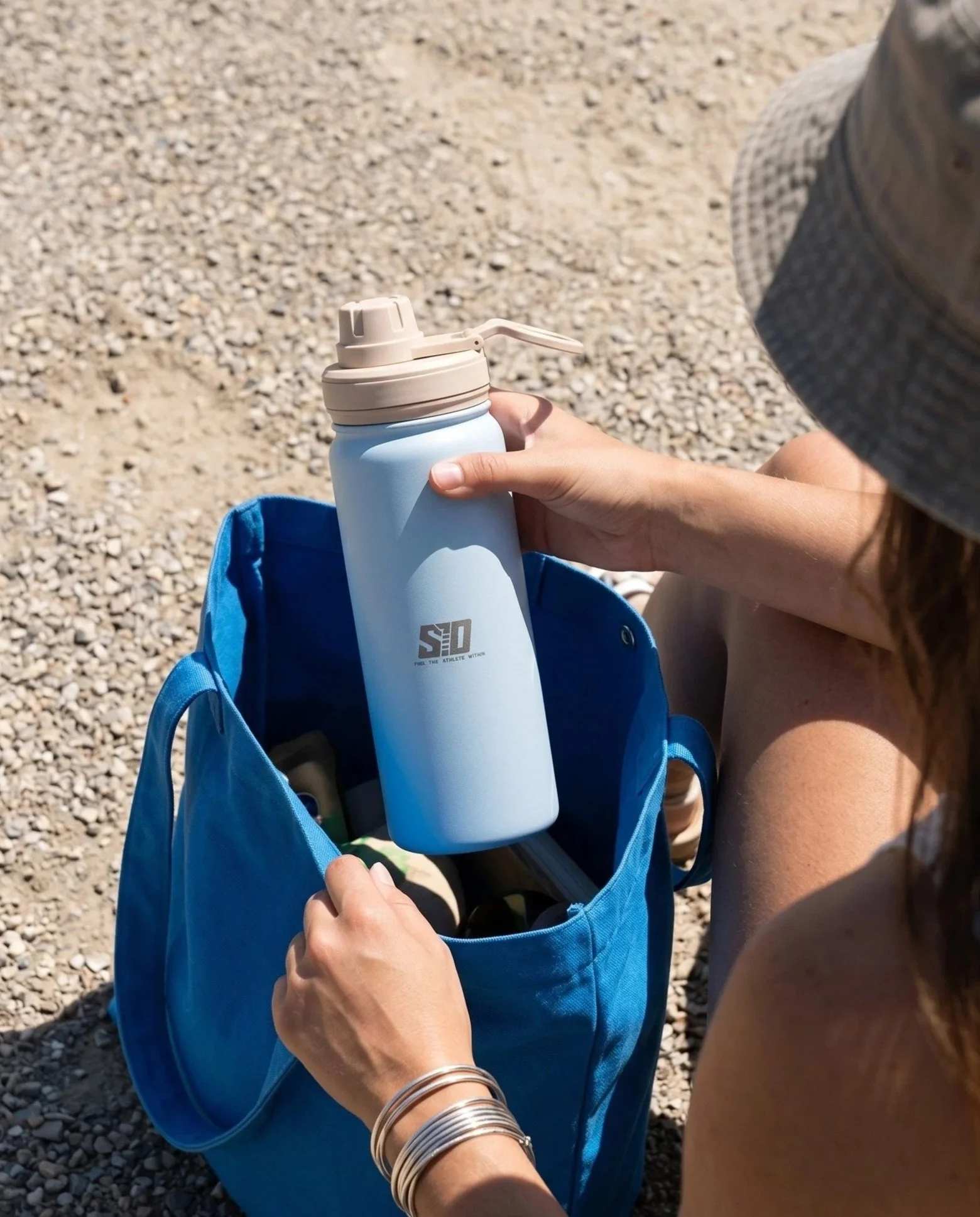 A person holding a light blue water bottle with a beige lid and spout, sitting on a pebble-covered ground, with a blue bag and wearing multiple silver bangles on their wrist.
