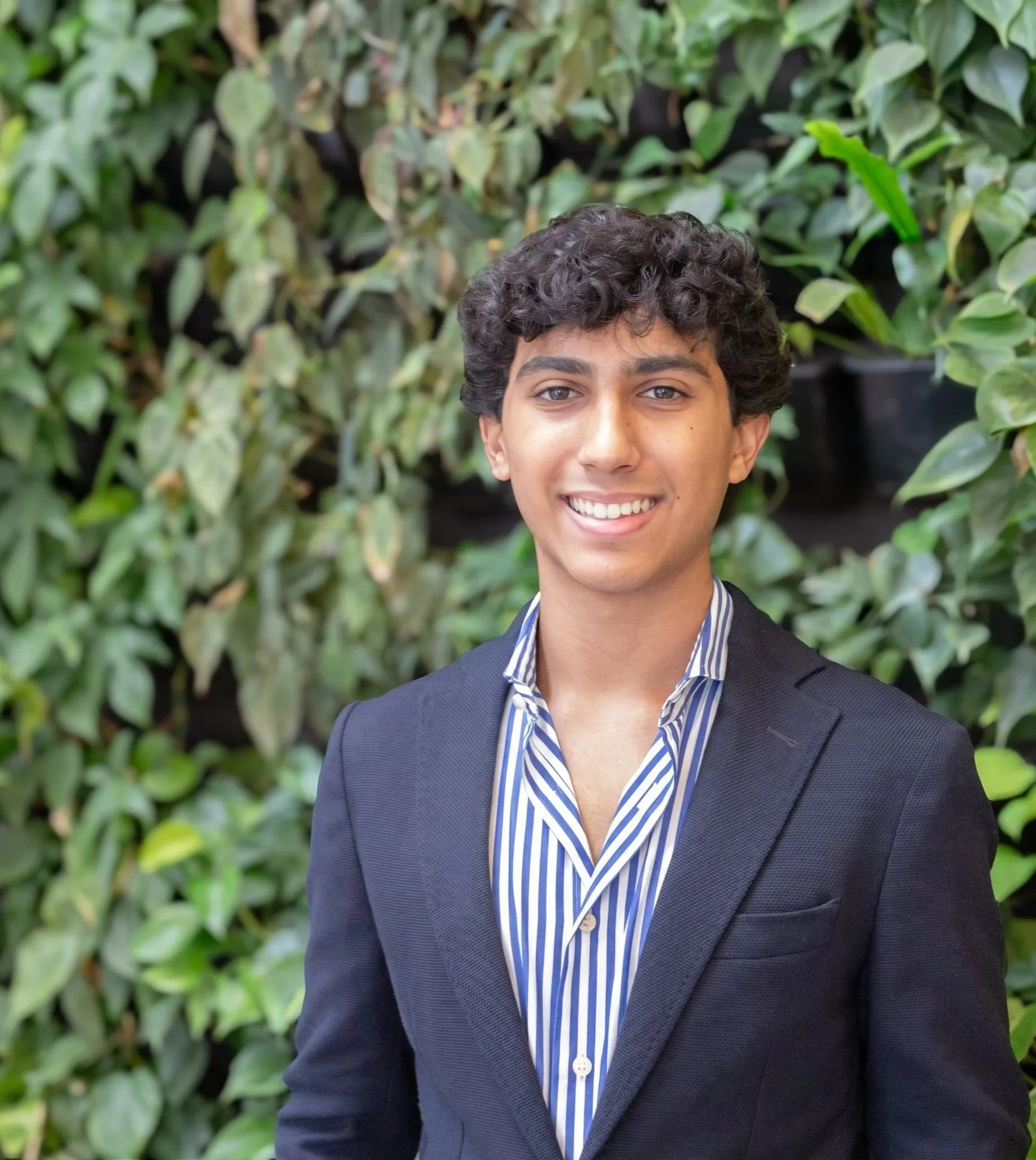 Portrait of a young man with dark curly hair, smiling, wearing a striped blue and white shirt and a dark blazer, standing in front of green leafy plants.