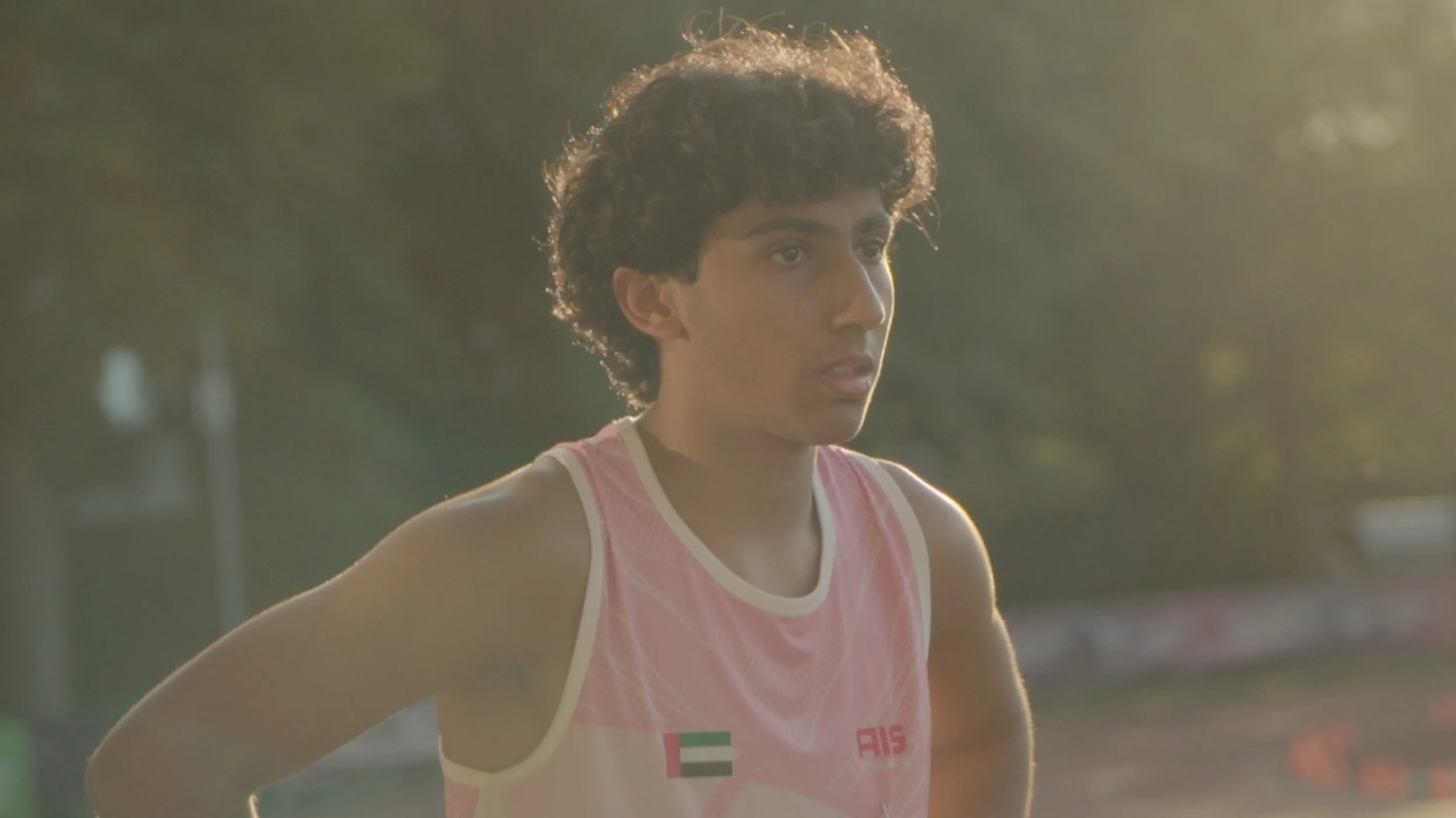 Young male athlete with curly hair wearing a pink and white sports jersey, standing outdoors during sunset, looking ahead with a focused expression.