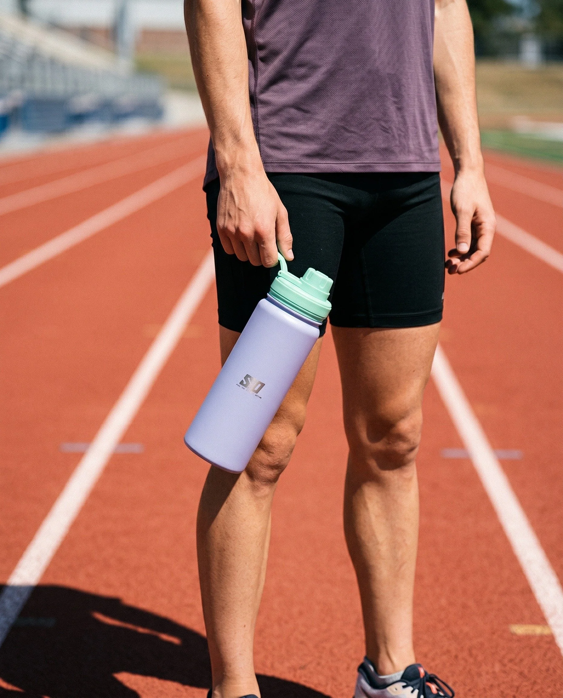 Person standing on running track holding a light purple water bottle, wearing black sports shorts, a purple shirt, and running shoes, with a background of a sports field and bleachers.