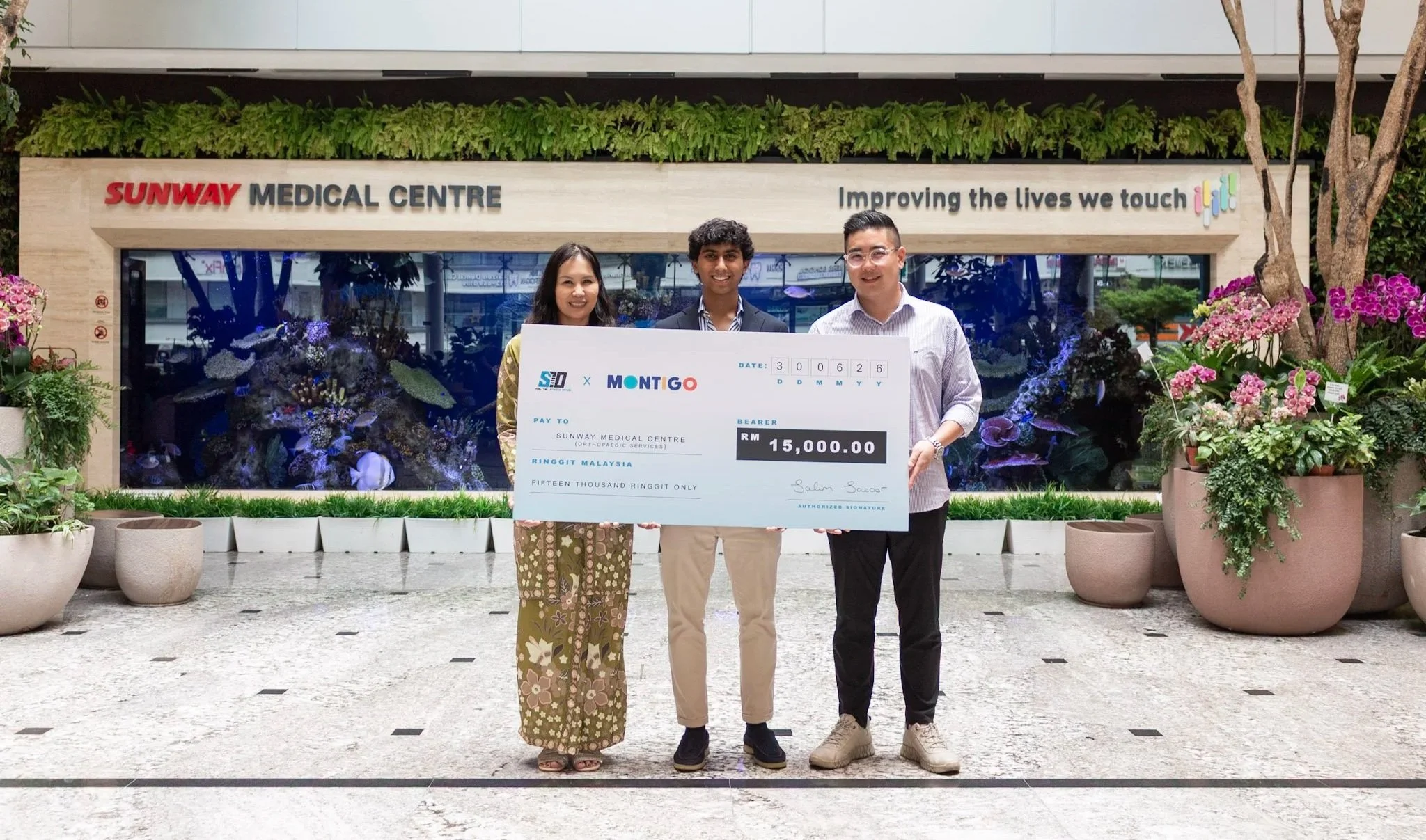 Three people standing outside a medical center holding a giant check for 15,000 Malaysian Ringgit. The background features the Sunway Medical Centre sign and greenery.